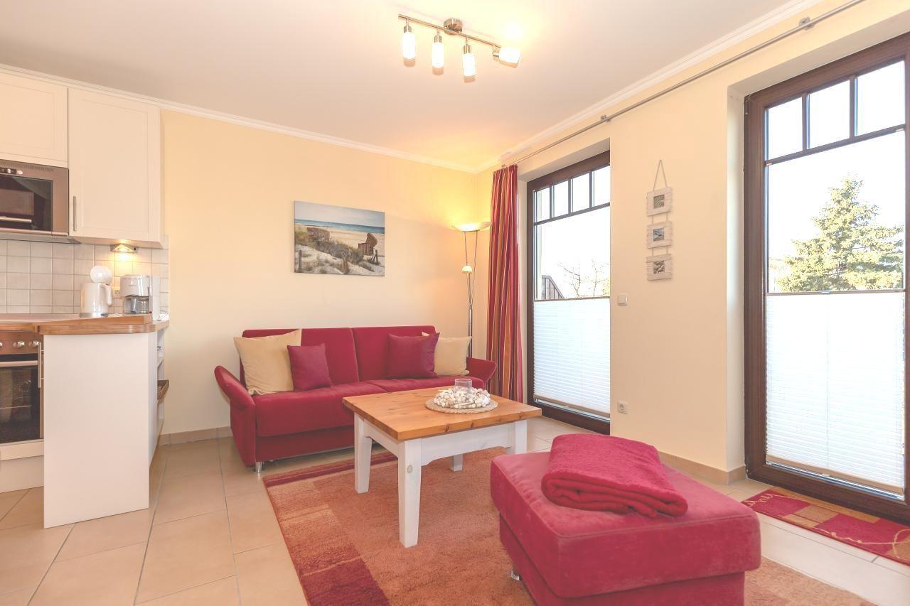 Wohnbereich mit rotem Sofa, Holztisch und Fenster. Küche mit Arbeitsplatte und Geräten.