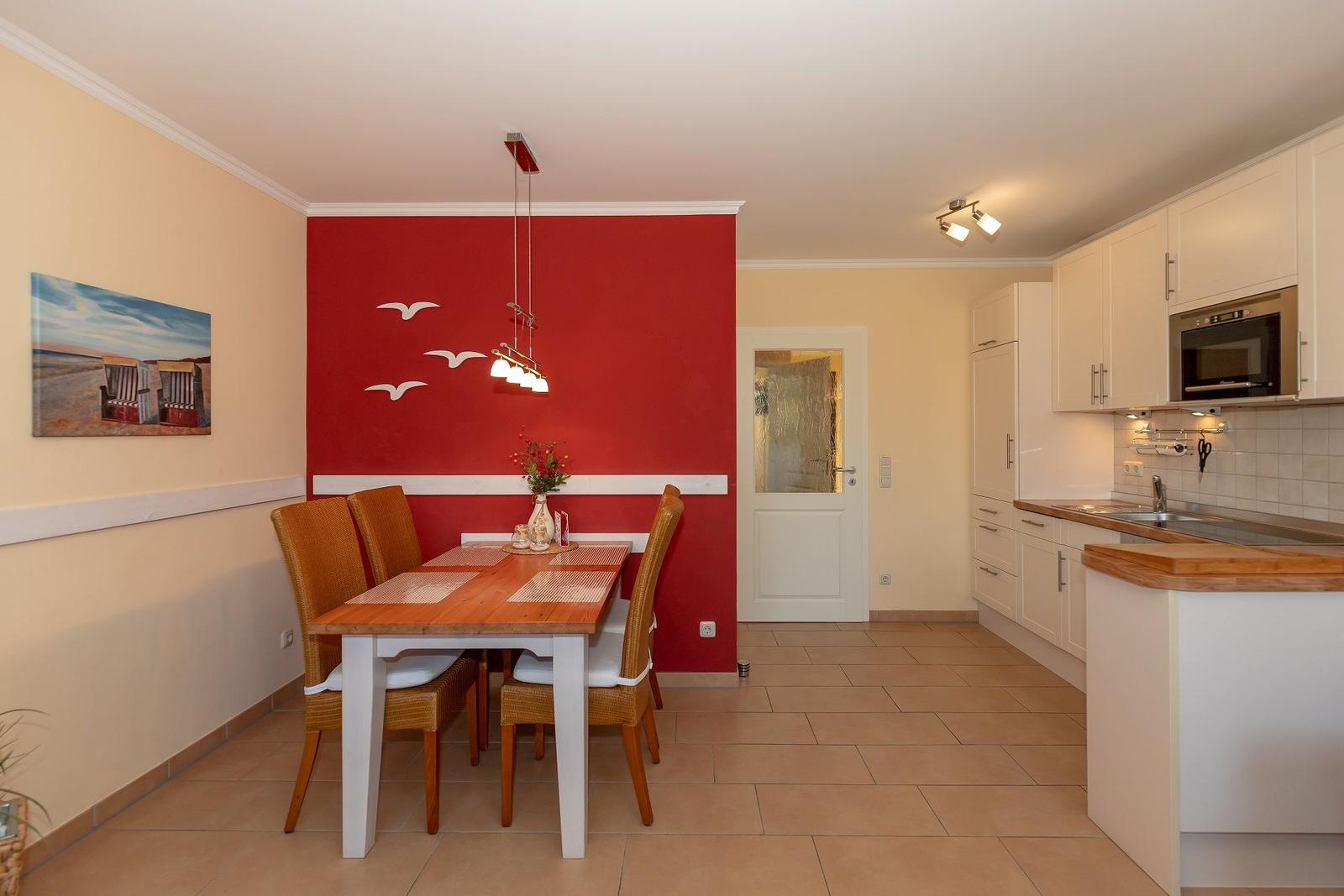 Essbereich mit Tisch und Stühlen, rote Wand mit Vögeln, Küche mit weißen Schränken und Holzoberfläche.
