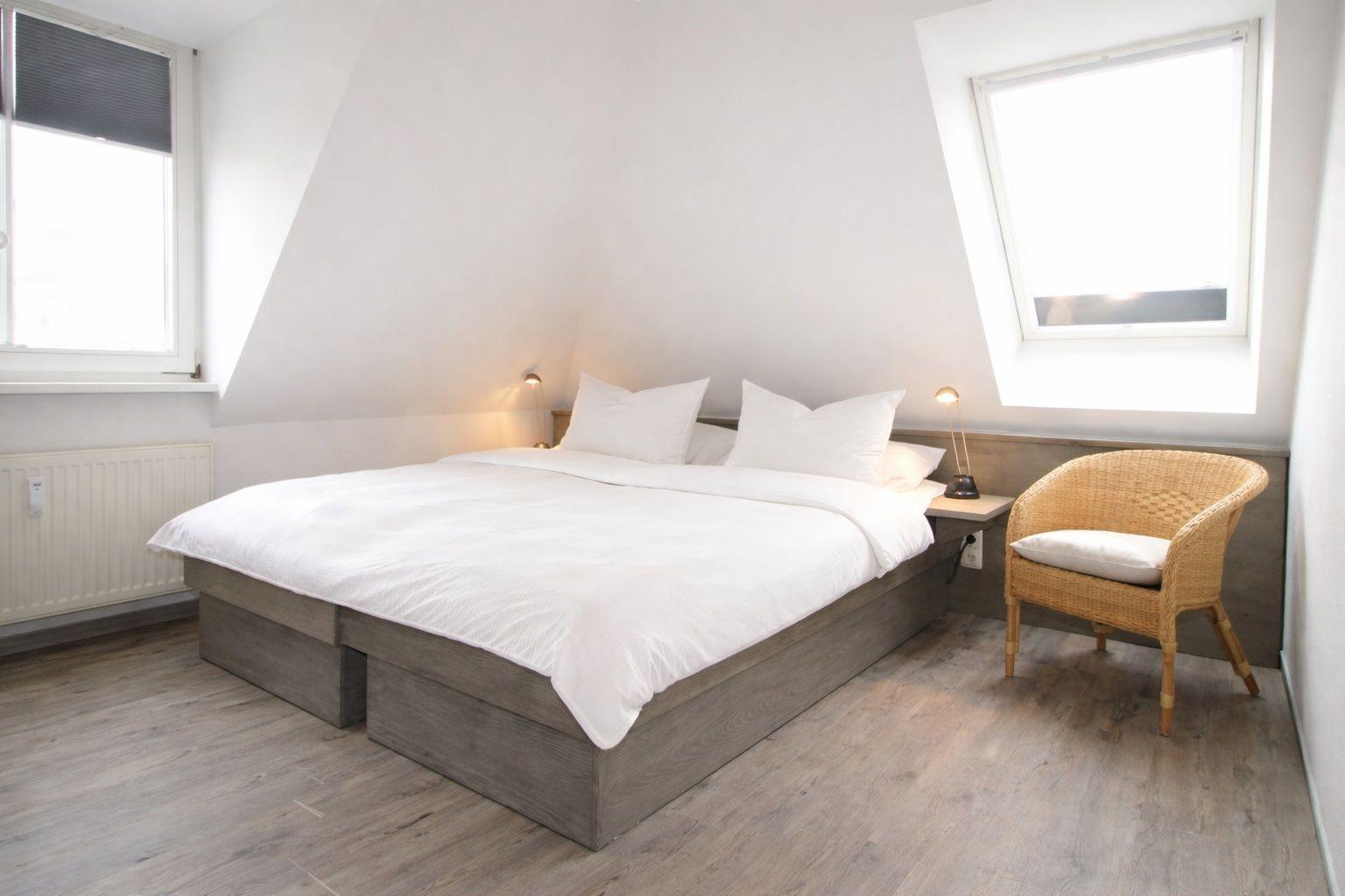 Schlafzimmer mit Doppelbett, Sessel und zwei Fenstern.