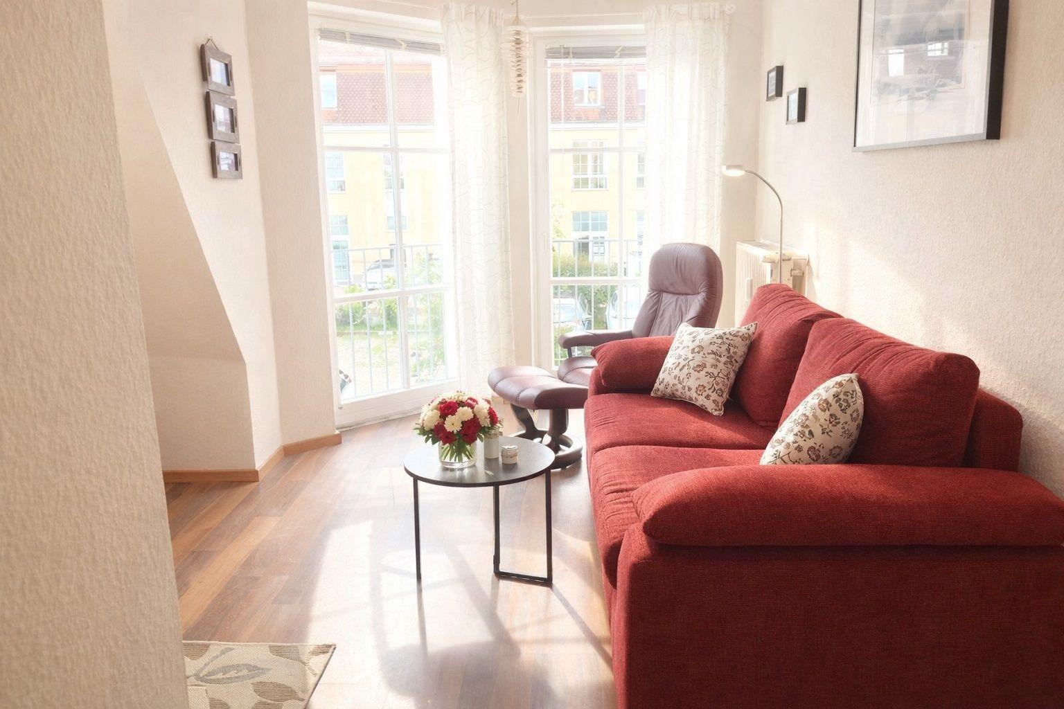 Ein Wohnzimmer mit rotem Sofa, Glasfront und Holzboden. Ein kleiner Tisch steht dazwischen.