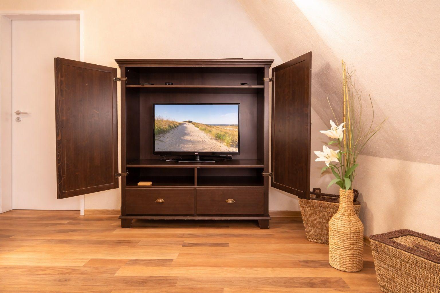 Ein dunkles Holz-TV-Möbel mit geöffneten Türen und einem Fernseher. Daneben steht ein geflochtener Vase mit Blumen.