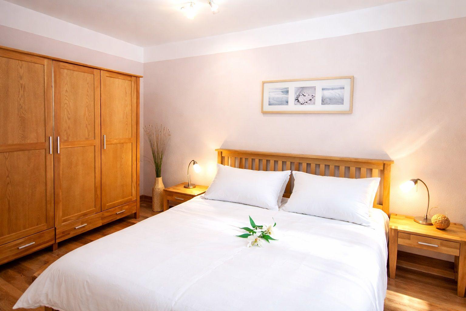 Schlafzimmer mit Holzbett, Nachttischen und Kleiderschrank. Bett mit weißen Kissen und Blumen.