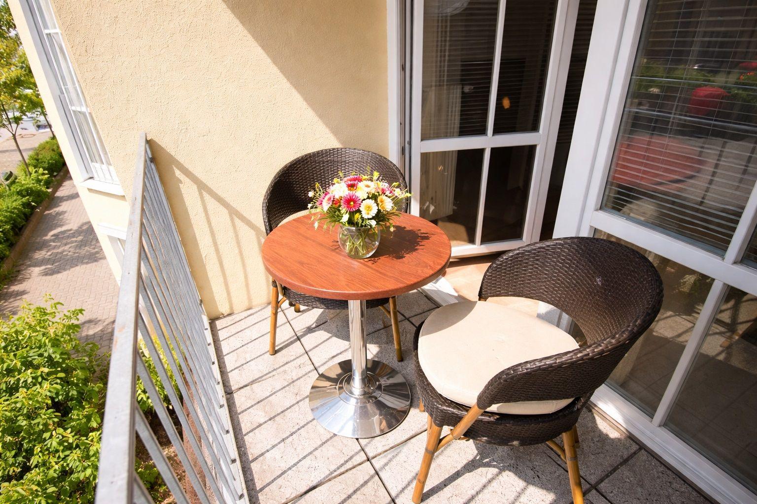 Balkon mit Tisch, zwei Stühlen und Blumenarrangement. Hintergrund: Hauswand und Fenster.