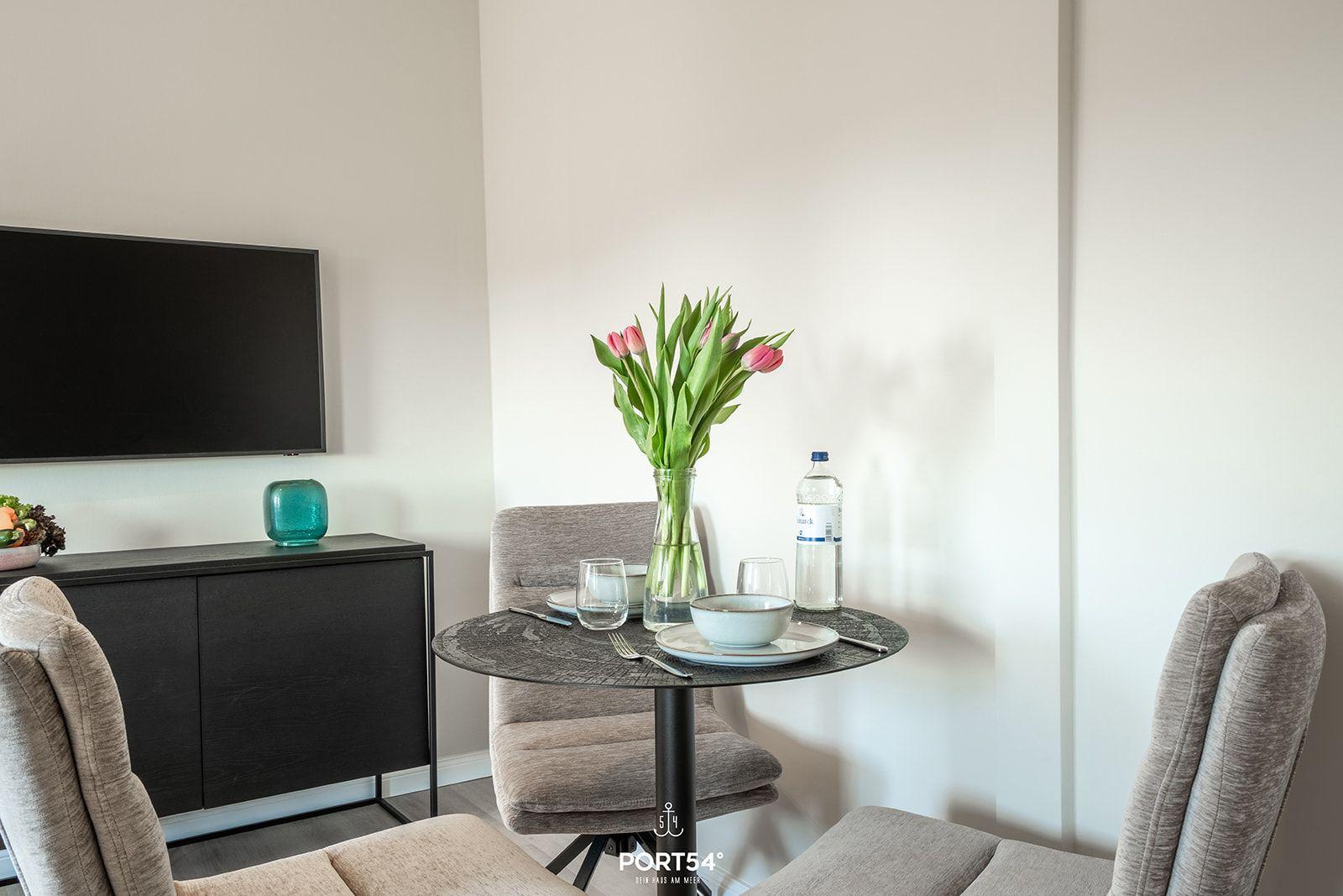 Dining table with chairs, flowers, and water bottle. TV and sideboard in background.