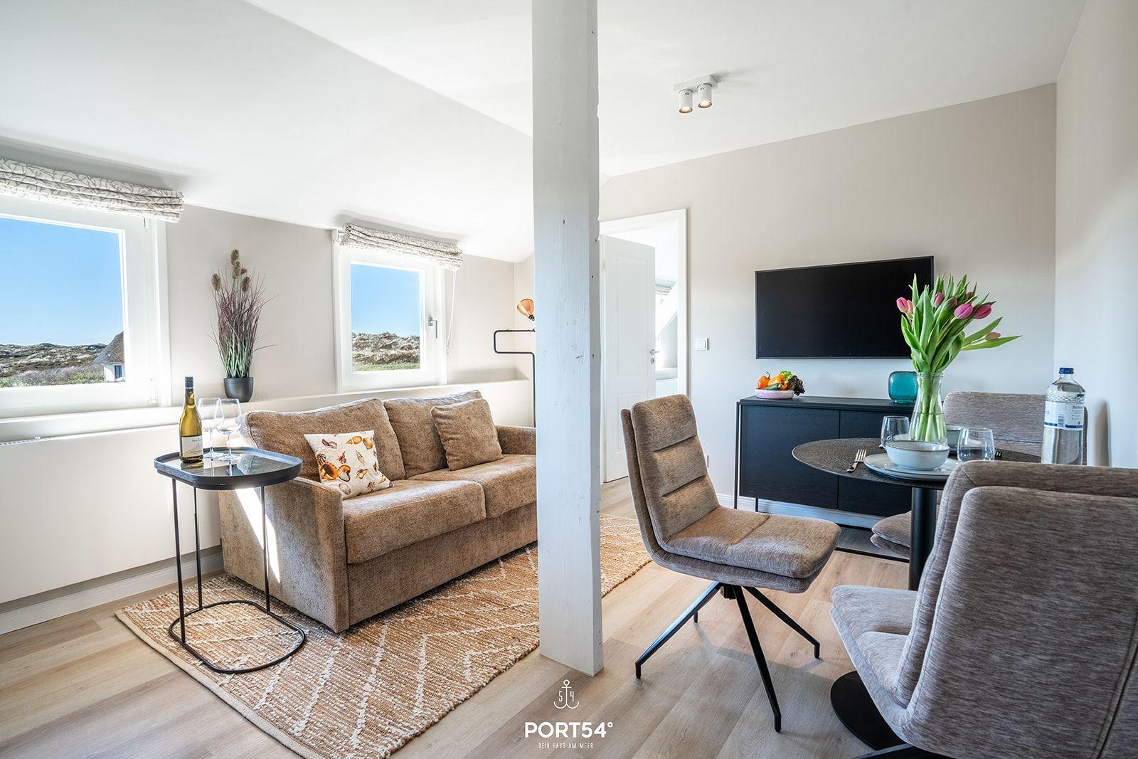 Living and dining area with sofa, TV, and table. Windows show landscape.