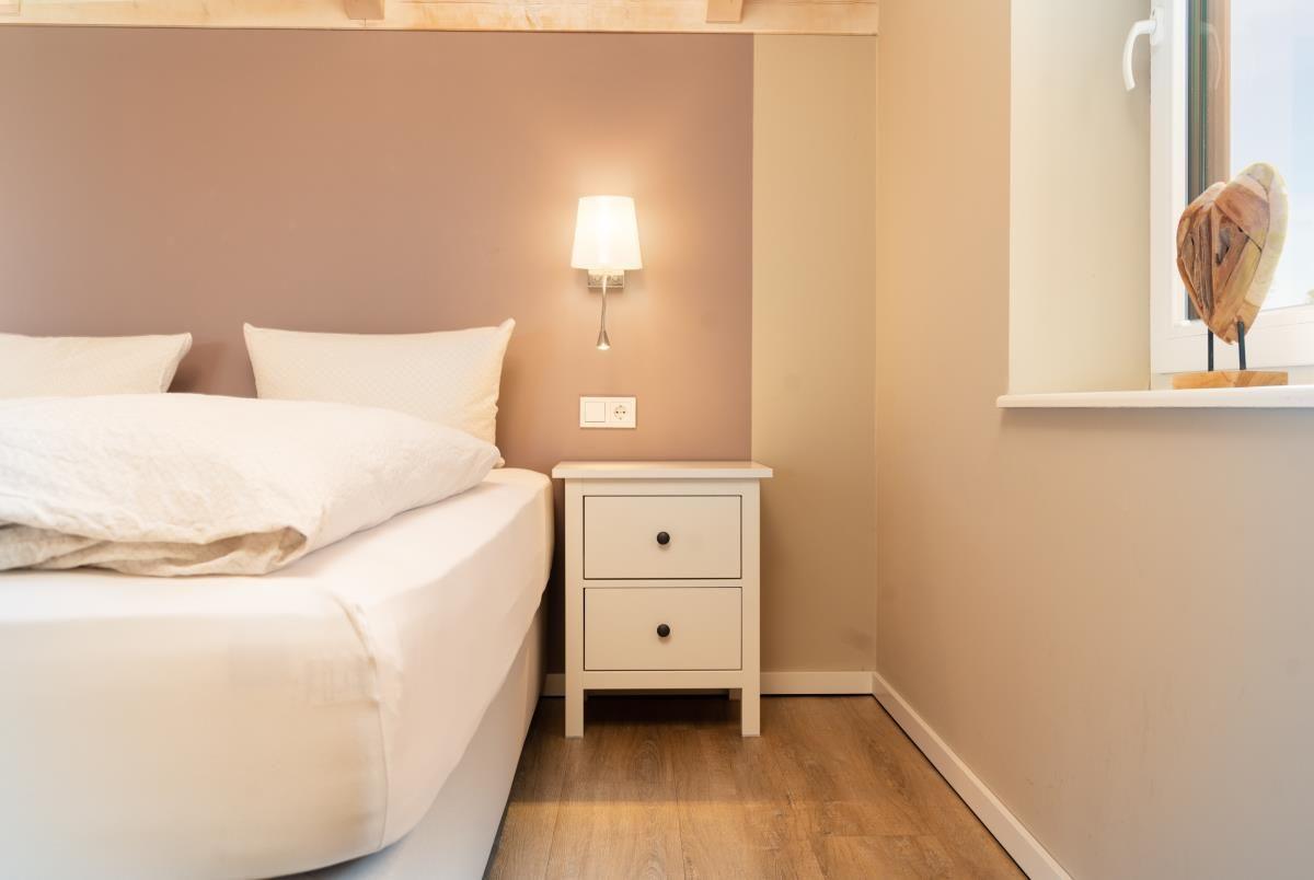 Schlafzimmer mit weißem Bett, Nachttisch und Wandlampe. Holzboden und Fenster mit Dekor.