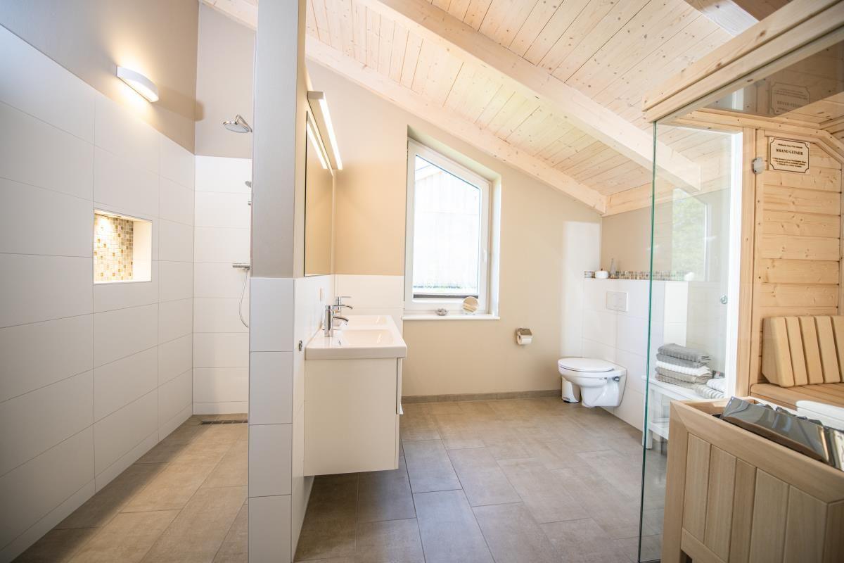 Badezimmer mit Dusche, Waschbecken und Toilette. Holzdecke und Glaswand zur Sauna.