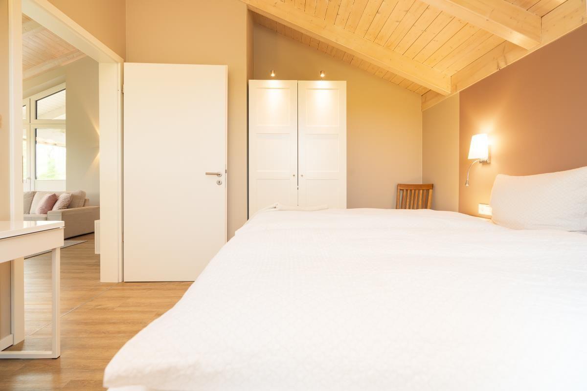 Schlafzimmer mit weißem Bett, Holzdecke und Tür zum Wohnbereich.