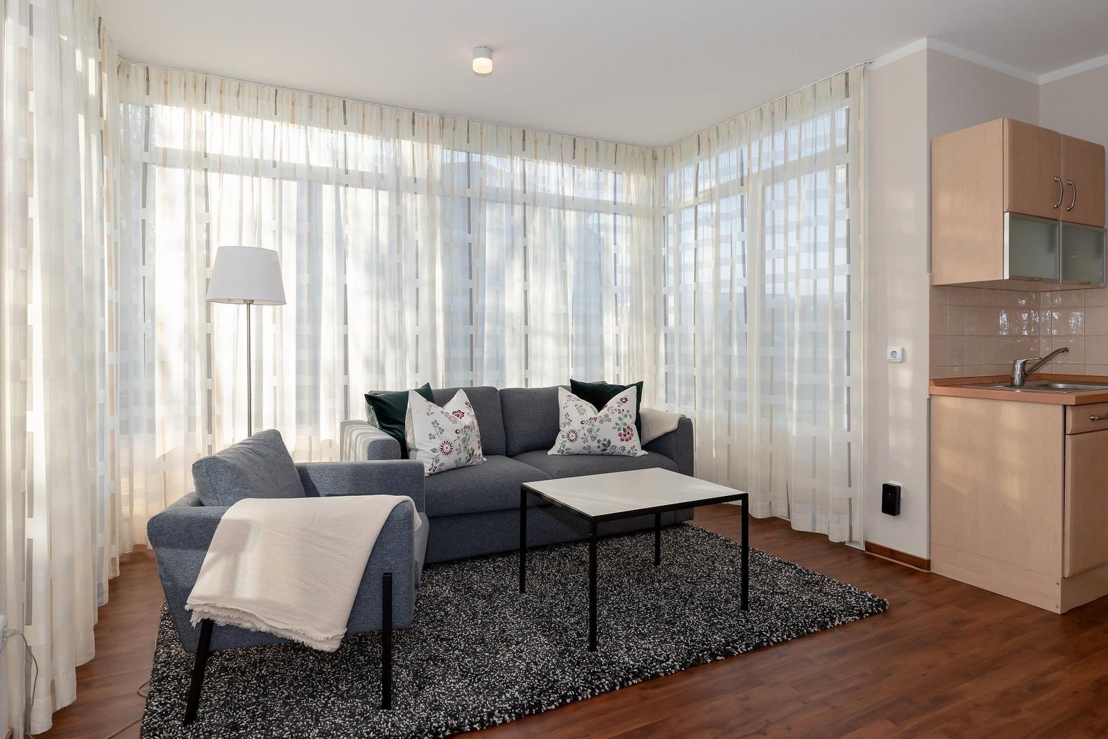 Wohnbereich mit Sofa, Couchtisch und Küchenzeile. Große Fenster mit durchsichtigen Vorhängen.