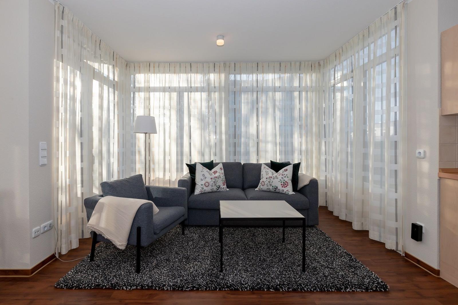 Wohnzimmer mit grauem Sofa, Tisch und Teppich vor großen Fenstern mit weißen Vorhängen.