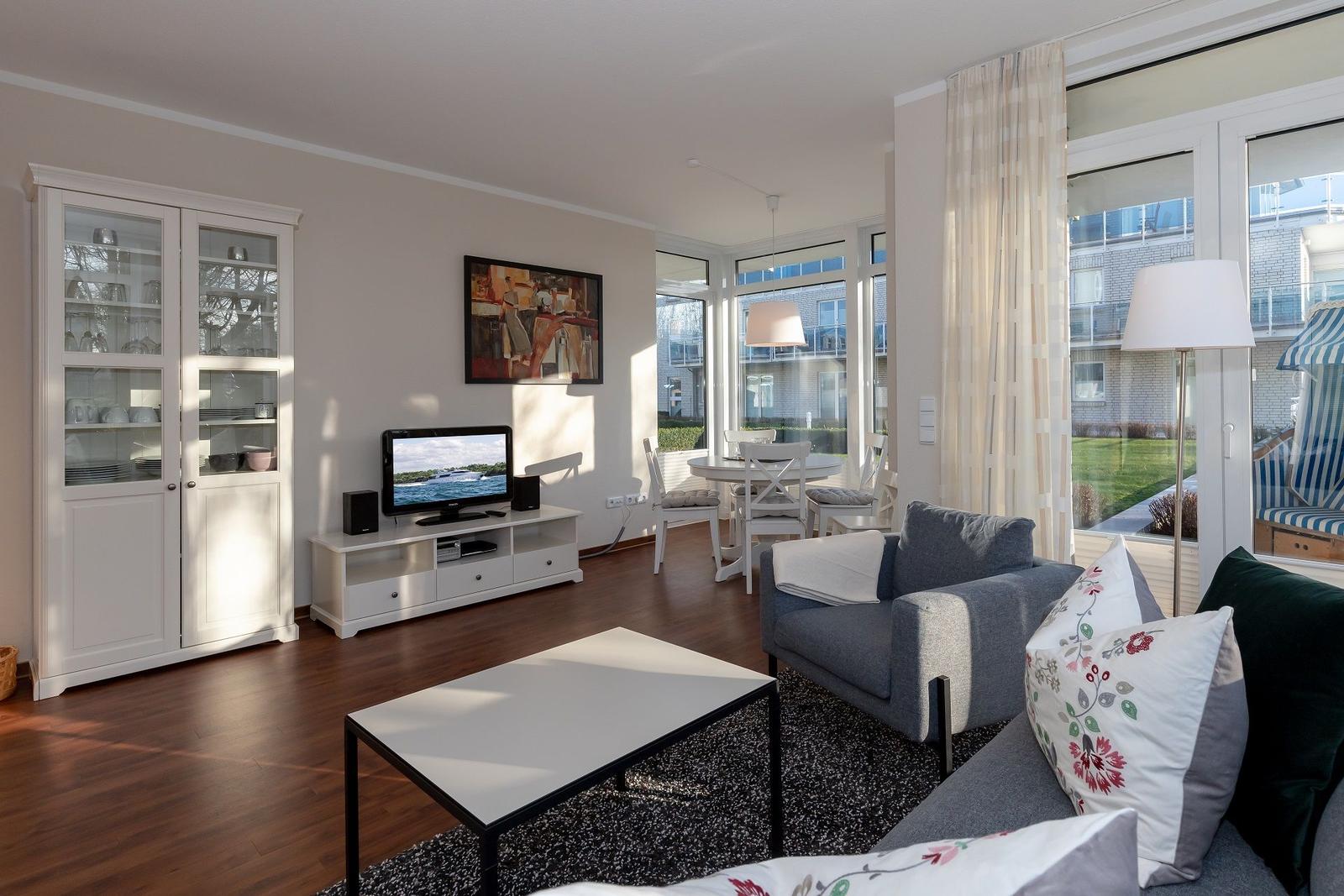 Wohnzimmer mit Sofa, Fernseher und Essbereich. Durchgang zum Balkon mit Gartenansicht.