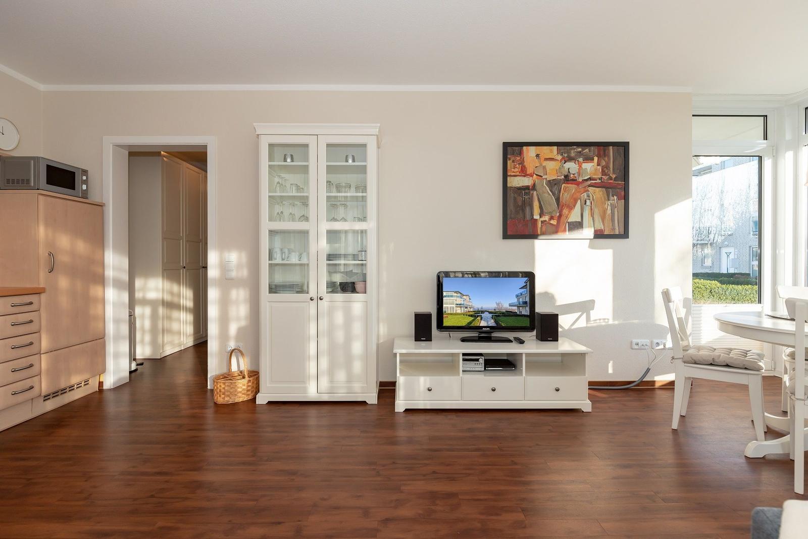 Wohnzimmer mit Fernseher, weißen Schränken und Holzboden. Fensterblick auf Garten.