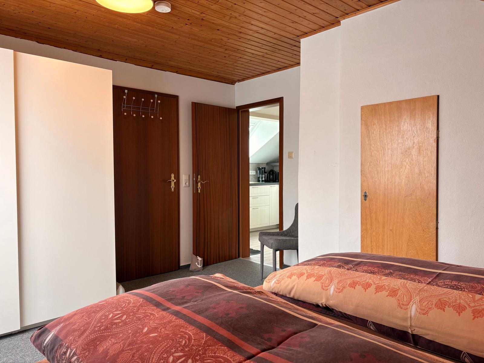 Schlafzimmer mit Bett, Holzdecke und Türen. Blick in die Küche.