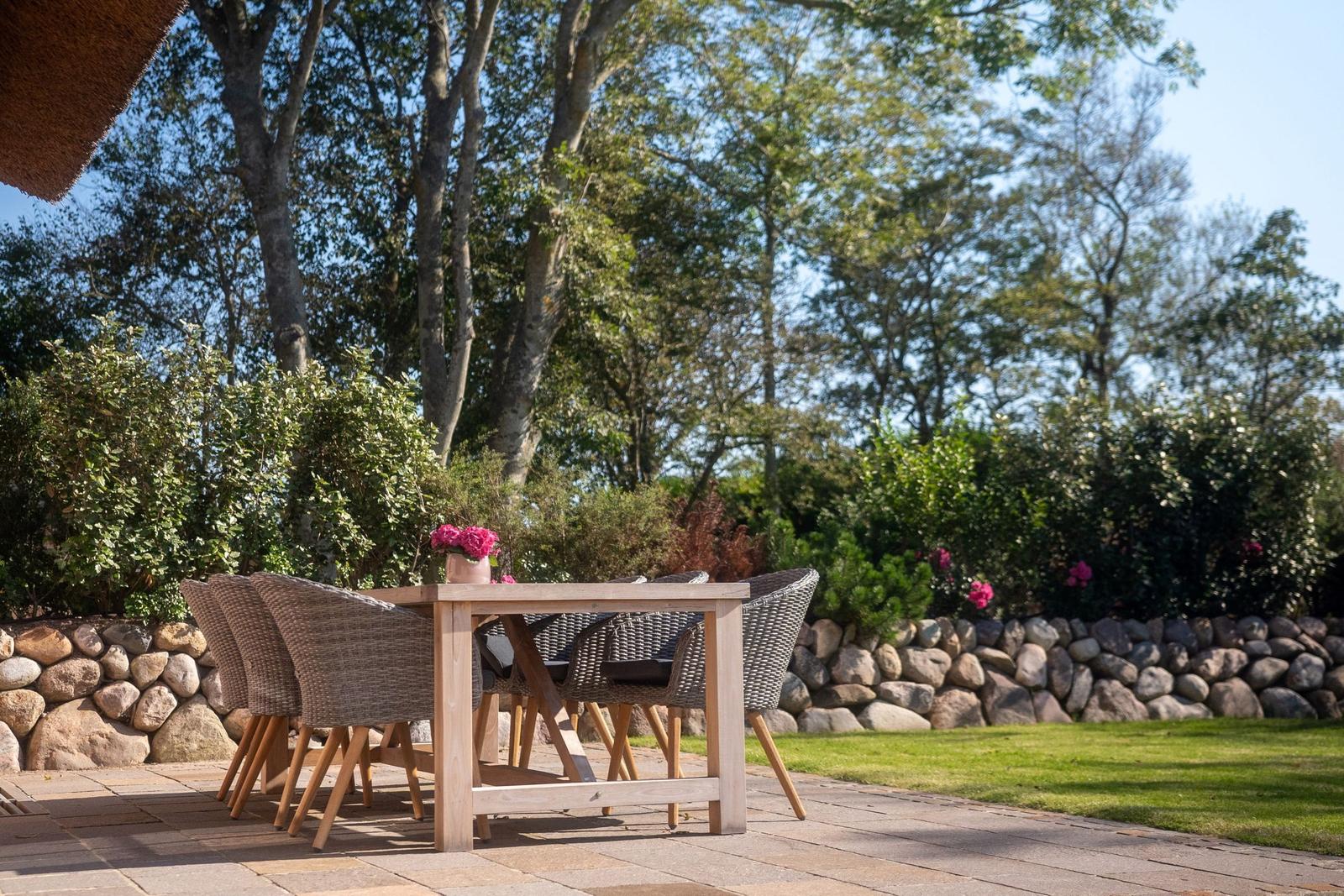 Holztisch mit Stühlen auf Terrasse, umgeben von Grün und Steinmauer.