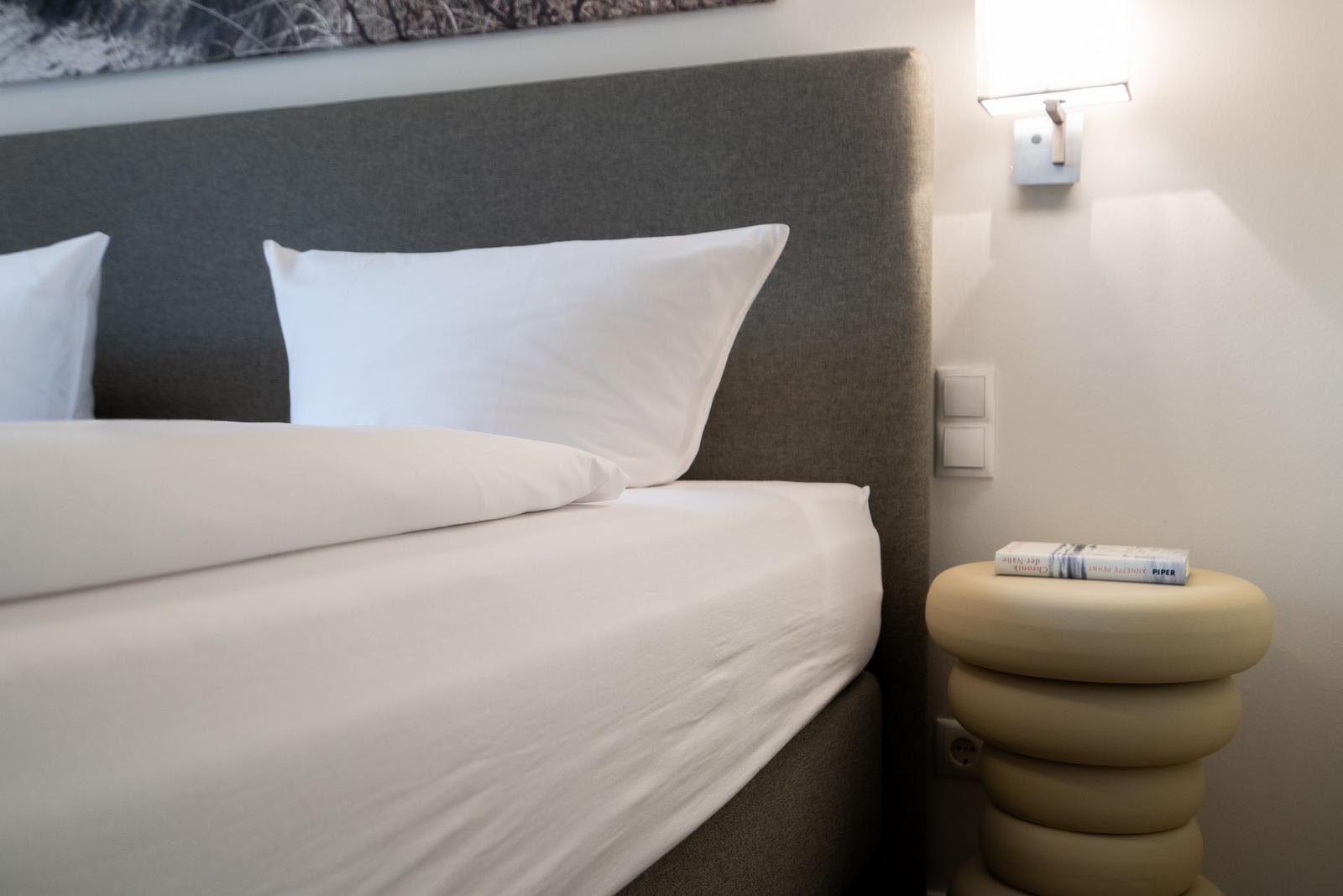 A bed with white bedding and pillows. Next to the bed is a round nightstand with a lamp.
