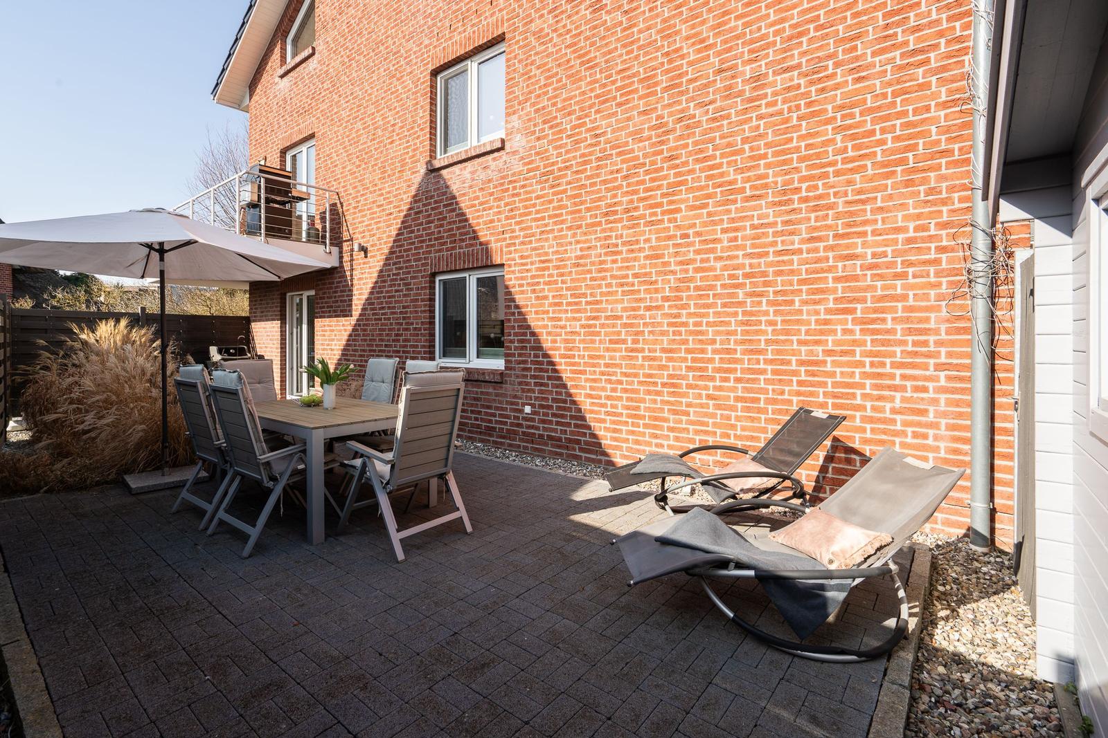 Terrace with table, chairs, and sun loungers in front of red brick house.
