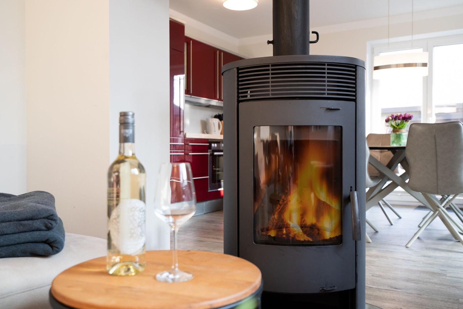 Fireplace burning, table with wine and glass. Kitchen and dining area in background.