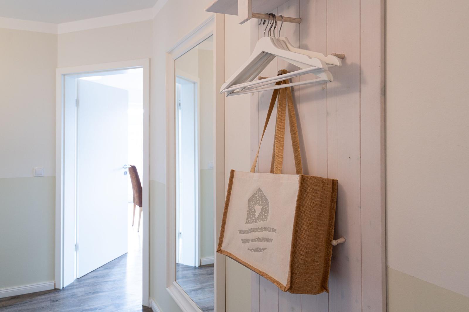 Hängende Tasche mit Holzständer an Wand, Spiegel und Tür im Hintergrund.