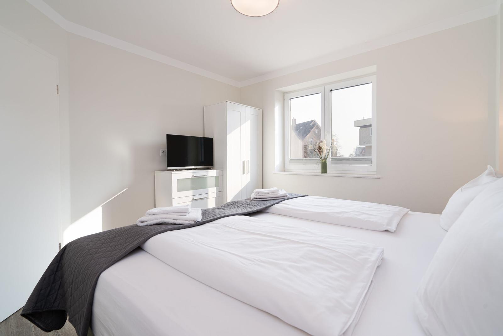 Schlafzimmer mit Bett, Fernseher und Fenster. Weiße Wände und Möbel.