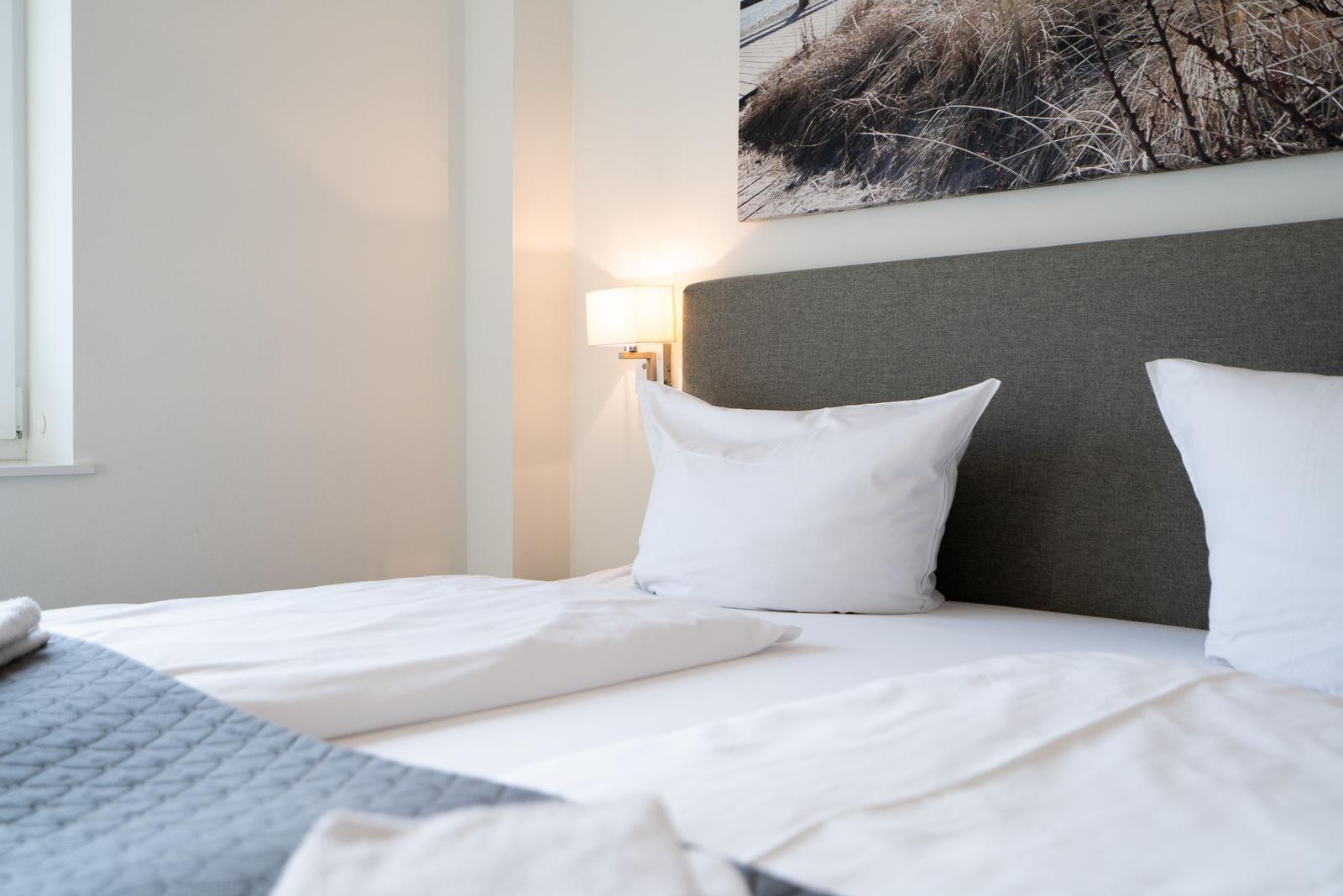 Doppelbett mit weißen Kissen und grauem Kopfteil. Wandbild und Nachttischlampe sind sichtbar.