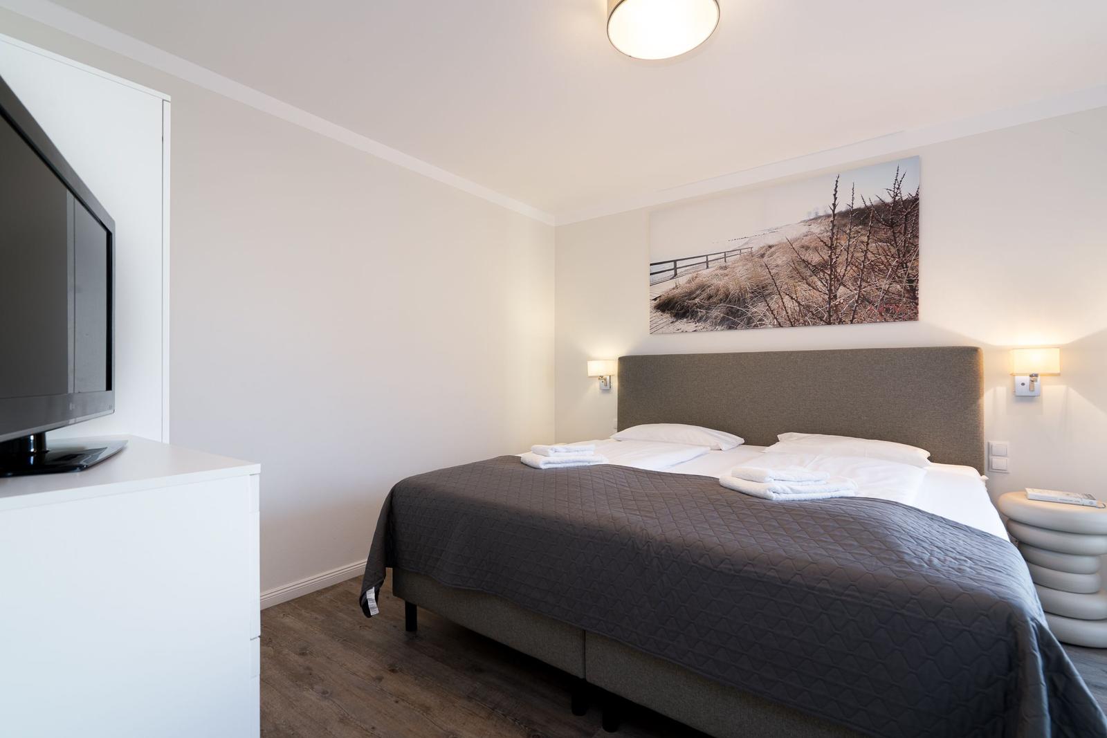 Schlafzimmer mit Bett, Wandbild und Fernseher auf weißem Schrank.