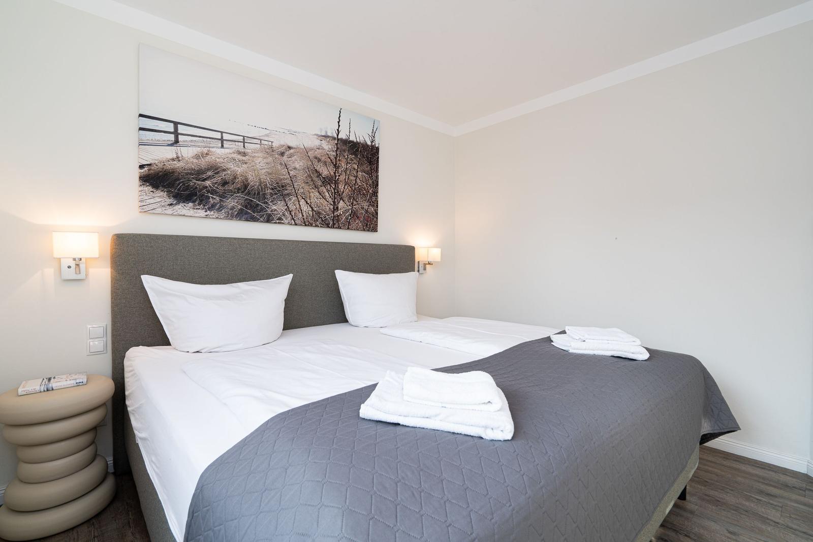 Ein Bett mit weißer Bettwäsche und grauer Decke. An der Wand hängt ein großes Bild.