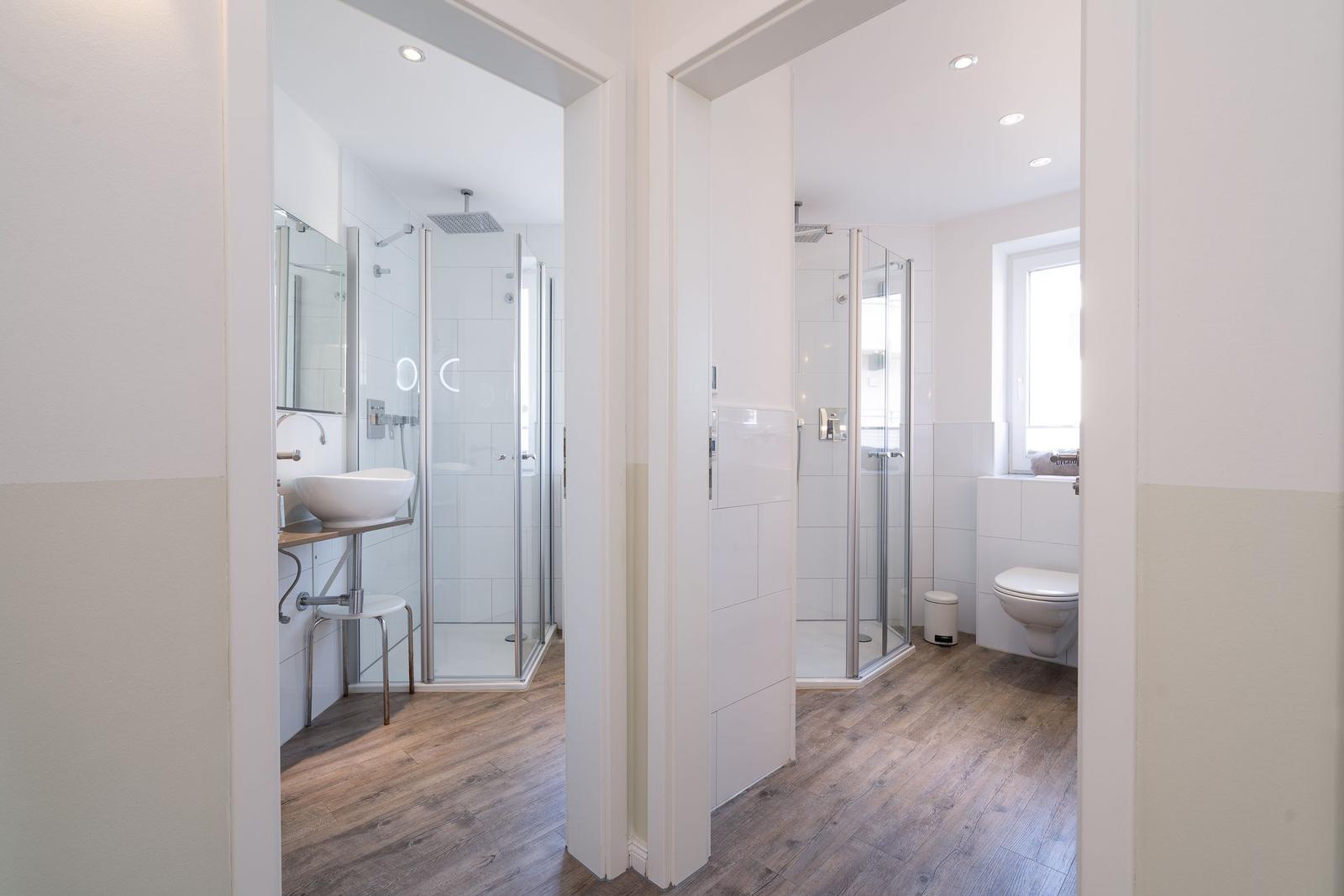 Zwei Badezimmer mit Duschen, Waschbecken und Toilette. Holzboden und weiße Wände.