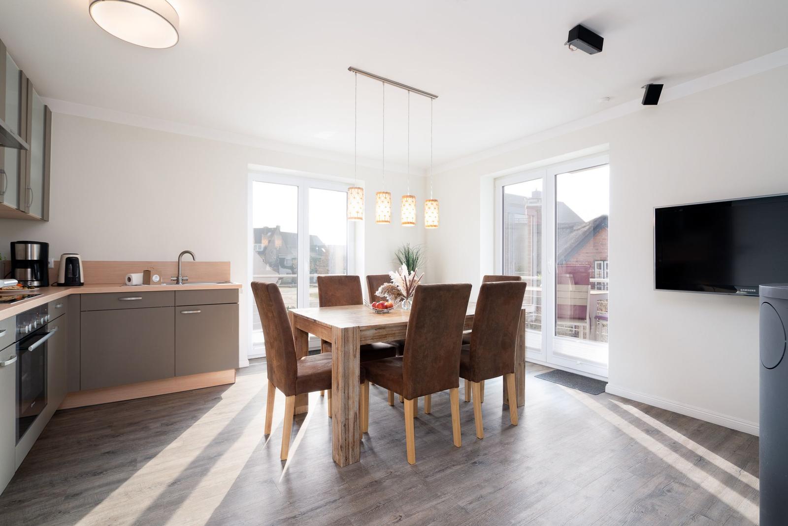 Essbereich mit Holztisch, sechs Stühlen und integrierter Küche. Große Fenster und TV-Wand.