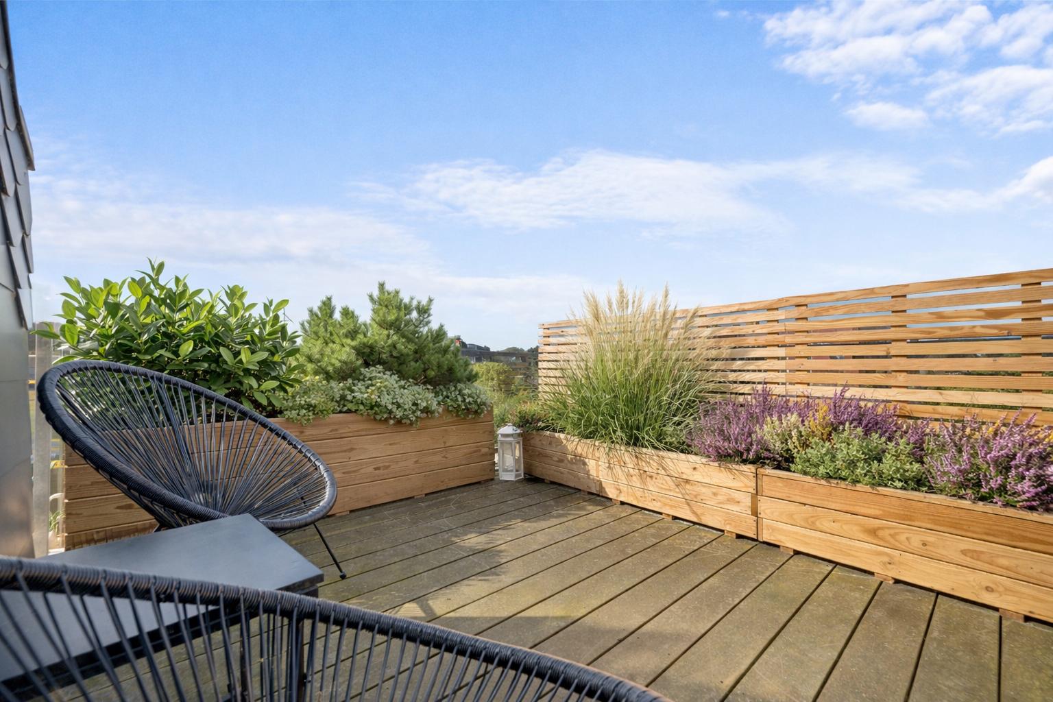 Terrasse mit Holzplanke, Pflanzenbeeten und Sitzmöbeln unter blauem Himmel.