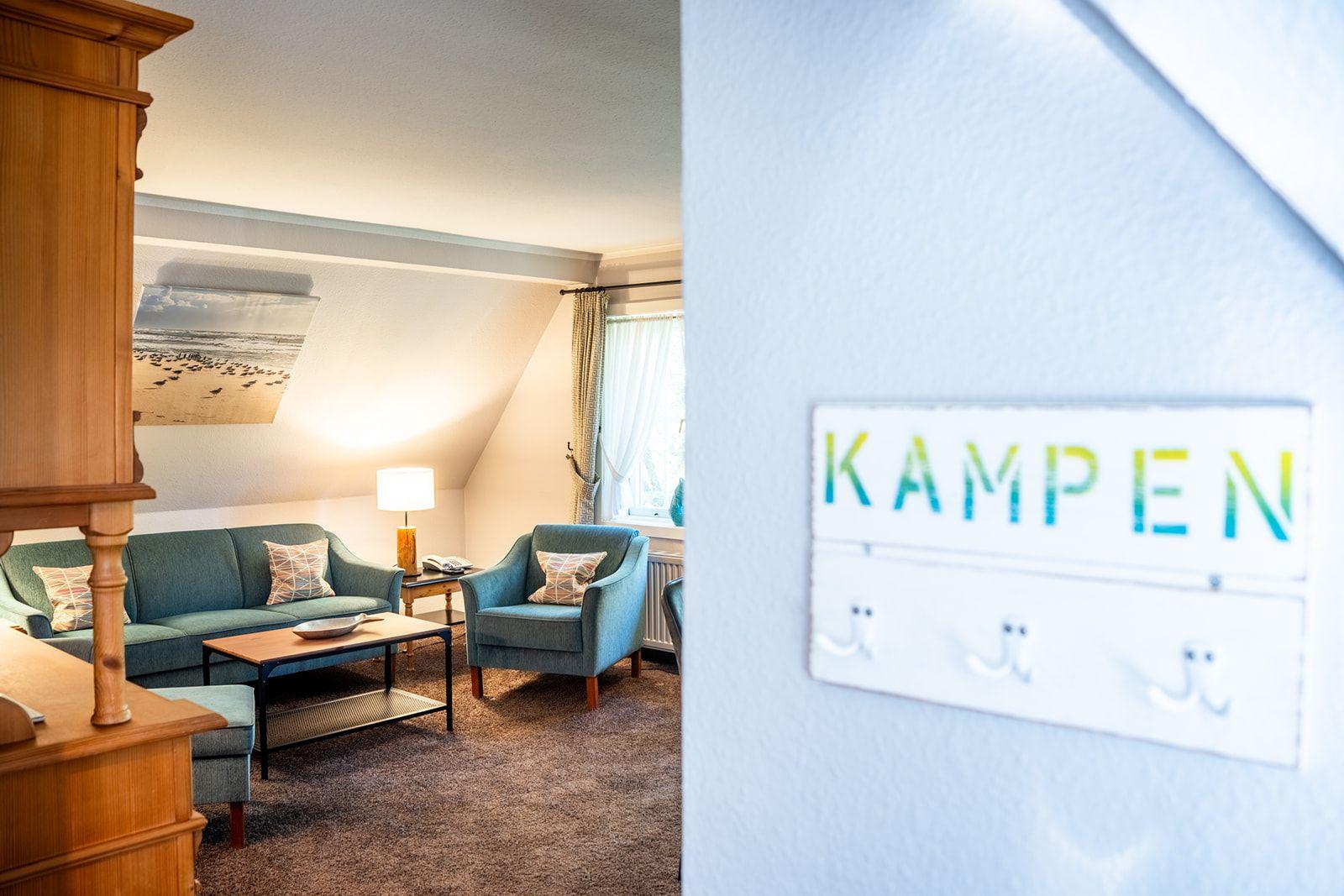 Wohnzimmer mit Sofa, Sessel und Tisch. Wandtafel mit KAMPEN.