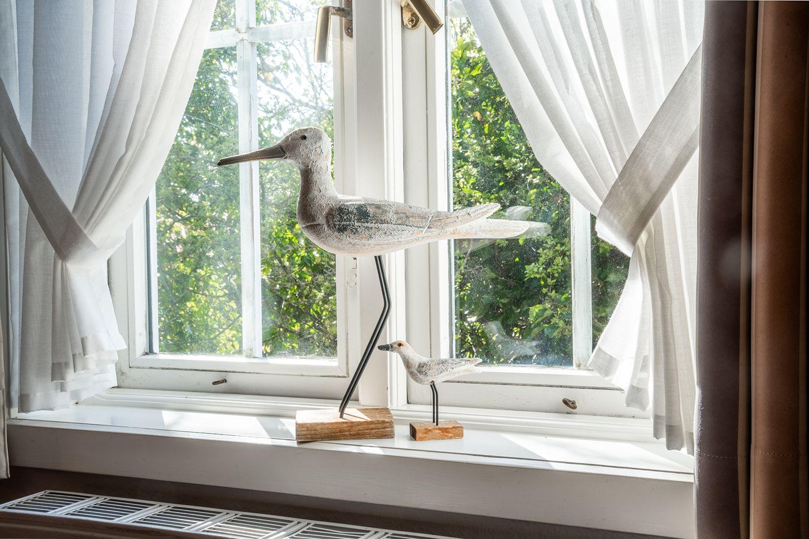 Zwei hölzerne Vogeldekorationen stehen auf dem Fensterbrett vor einem Fenster mit weißen Vorhängen.