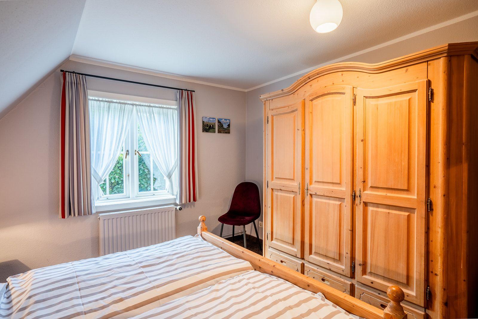 Schlafzimmer mit Holzschrank, Bett und Fenster mit Gardinen.