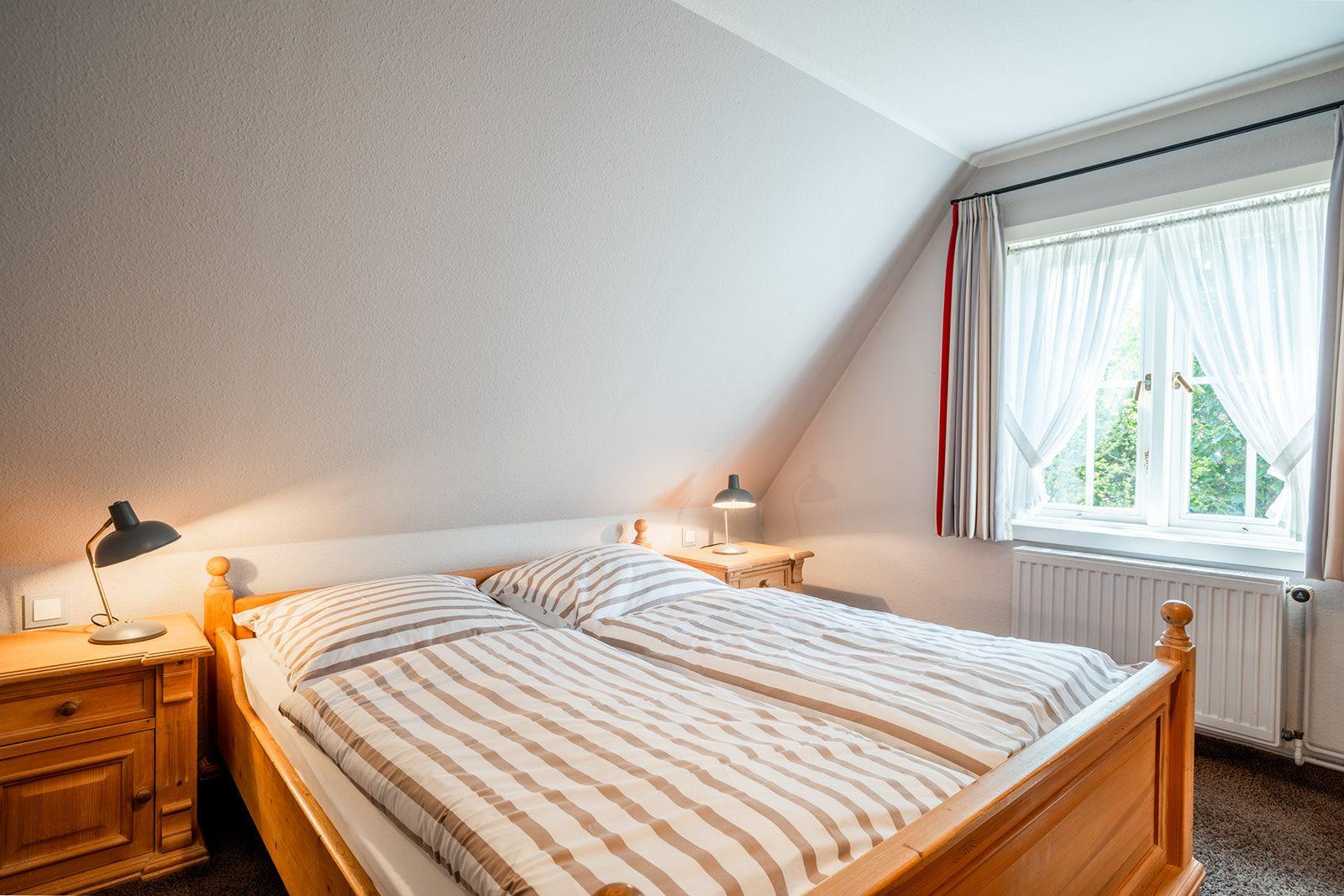 Doppelbett mit Streifenbettenlaken, zwei Nachttische mit Lampen und Fenster mit Vorhängen.