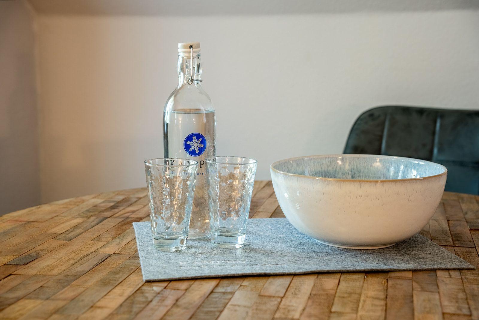 Holztisch mit Wasserglas, Flasche und Keramikschale.