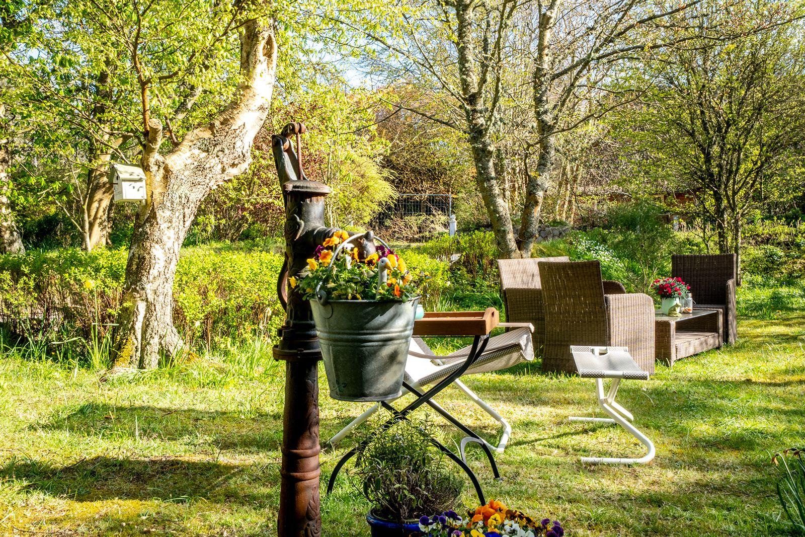 Garten mit Sitzgruppe, Blumen und altem Brunnen unter Bäumen.