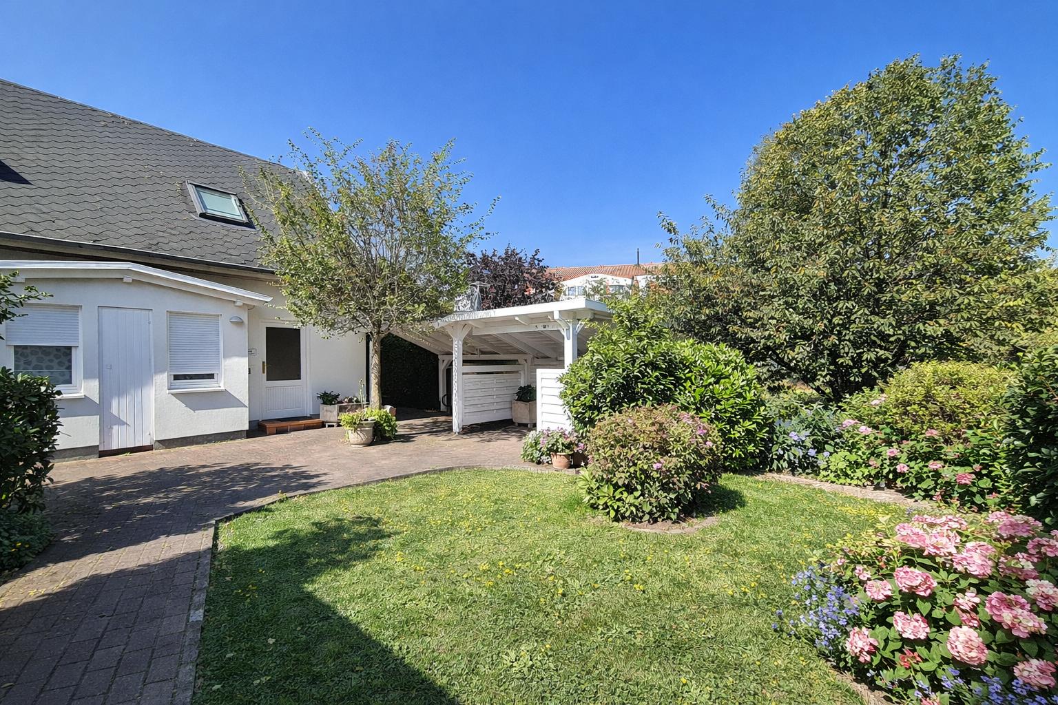 Haus mit Garten, Carport und Blumenbeeten unter blauem Himmel.