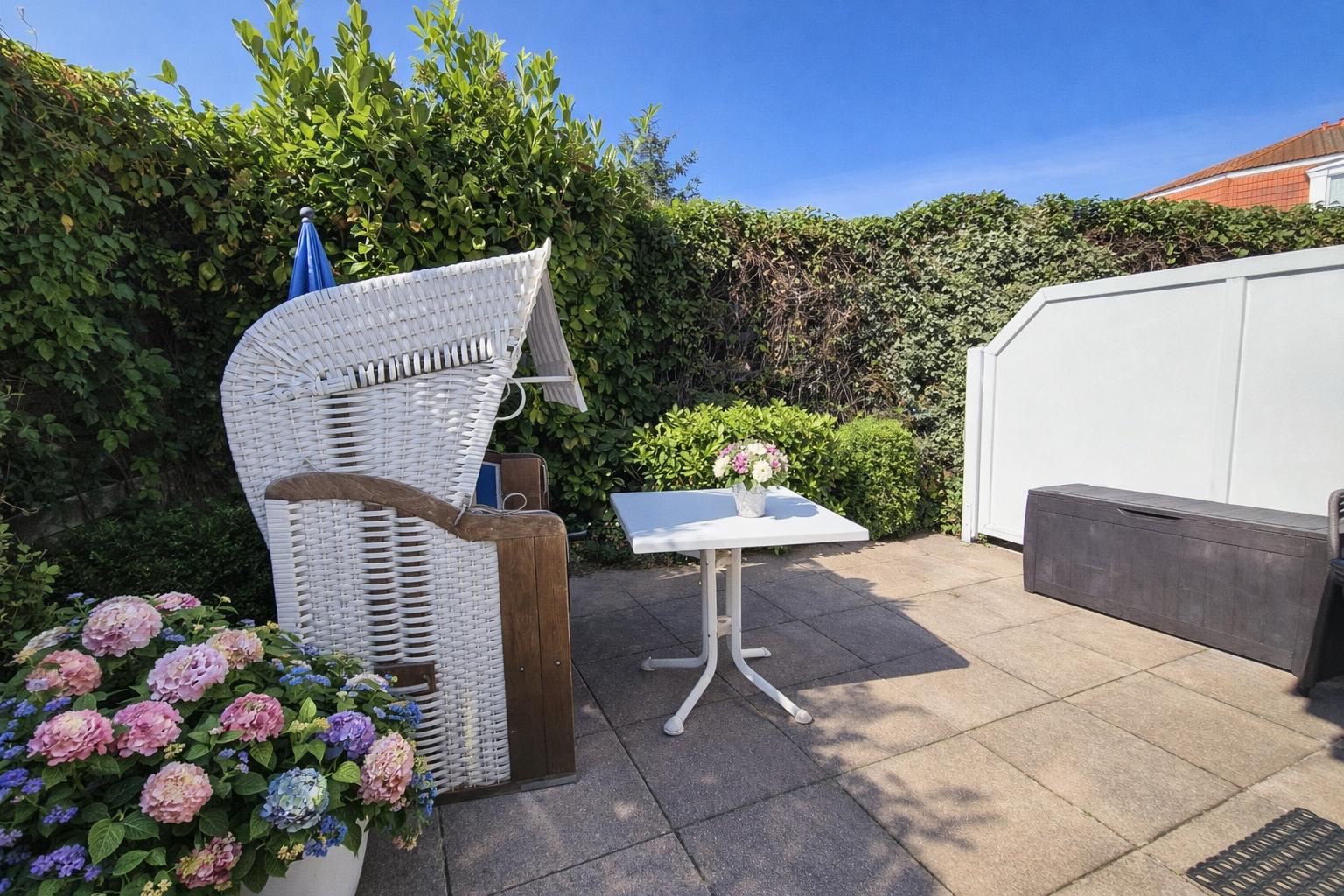 Terrasse mit weißem Stuhl, Tisch und Blumenbeet. Hintergrund: grüne Hecke und weißer Zaun.