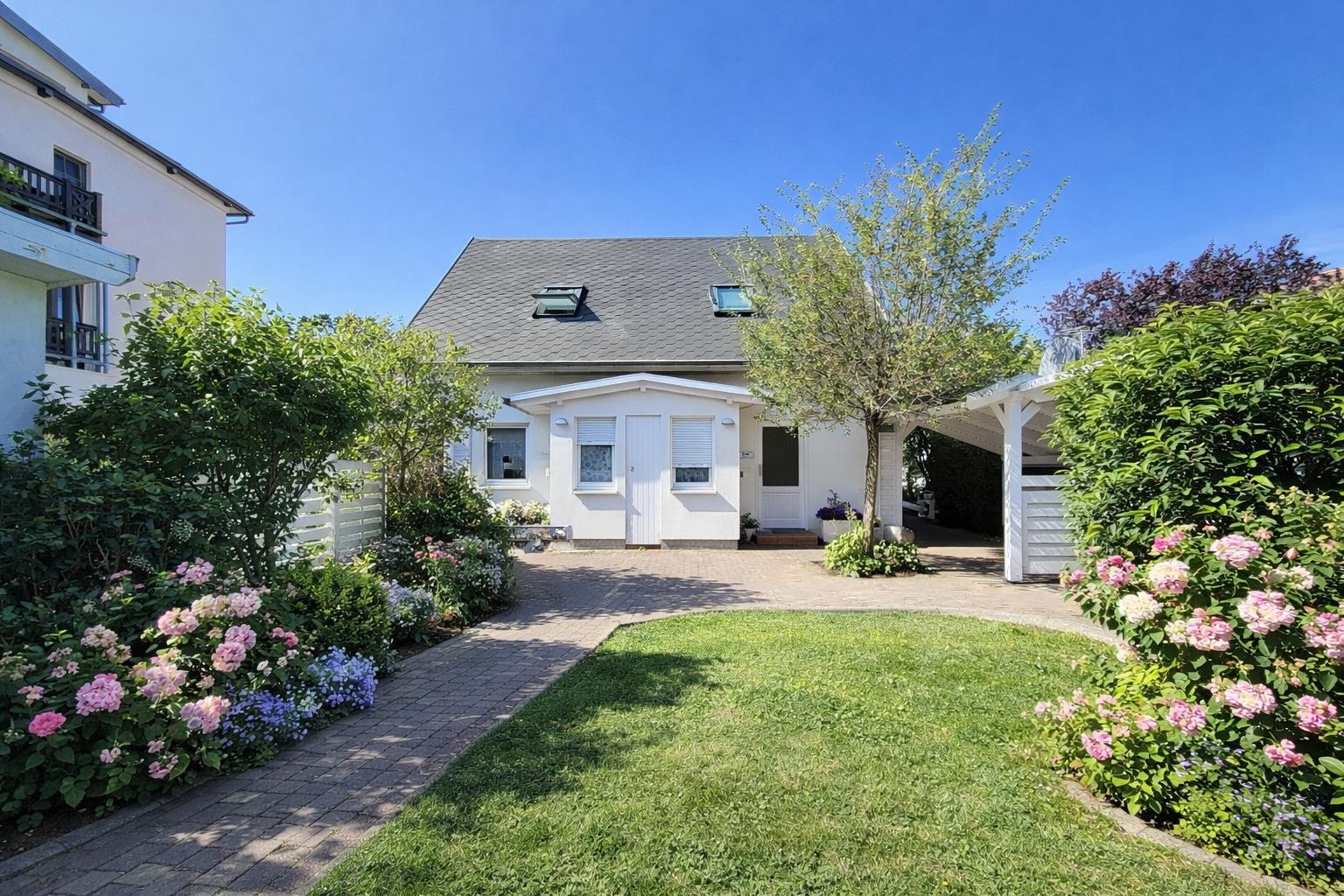 Weißes Haus mit Garten, Blumenbeete und Terrasse unter blauem Himmel.