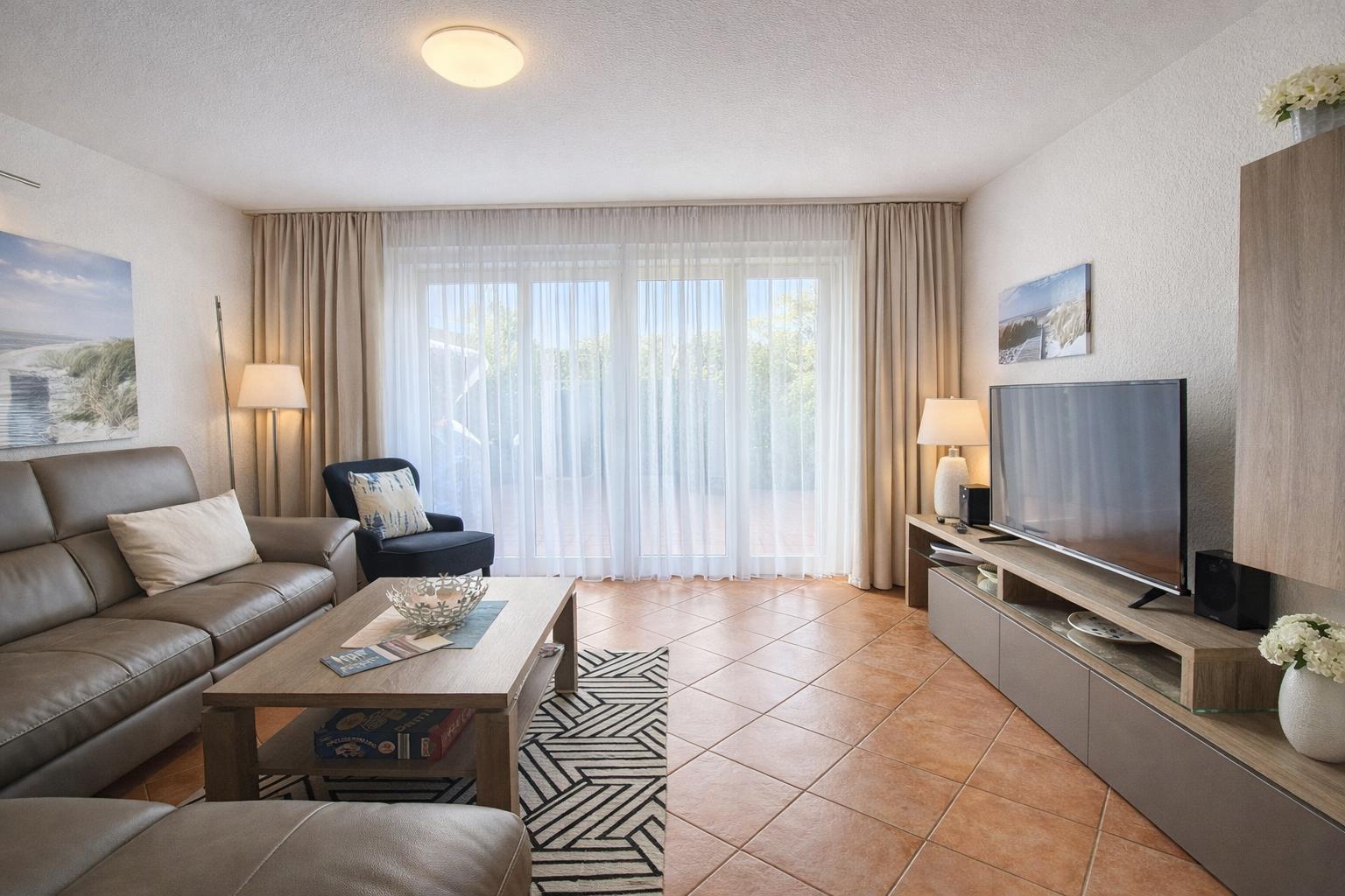 Wohnzimmer mit Sofa, Couchtisch und TV. Große Fensterfront mit Vorhängen.