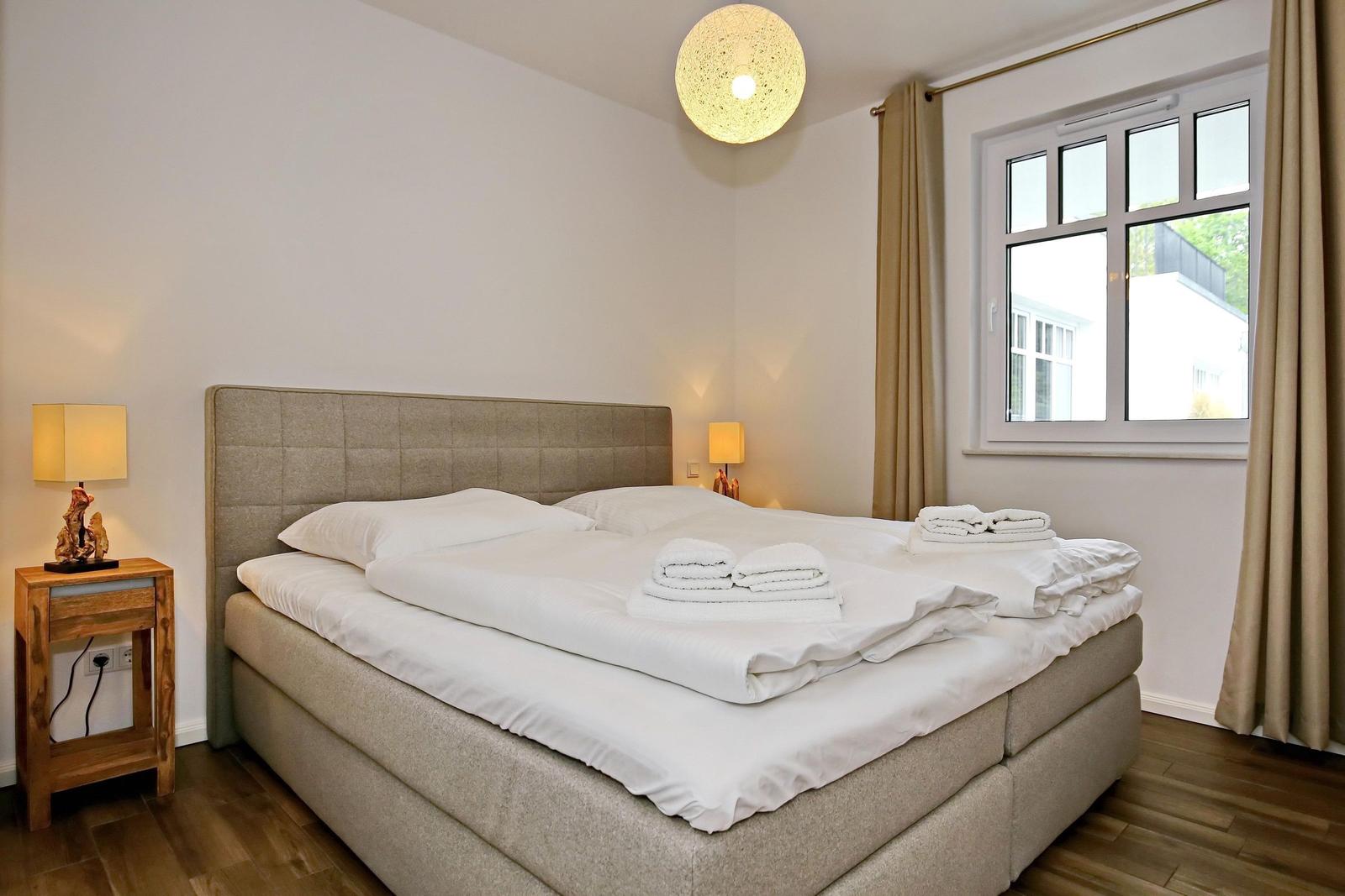 Doppelbett mit weißen Bettwäsche, Holzsideboard und Fenster mit Vorhängen.