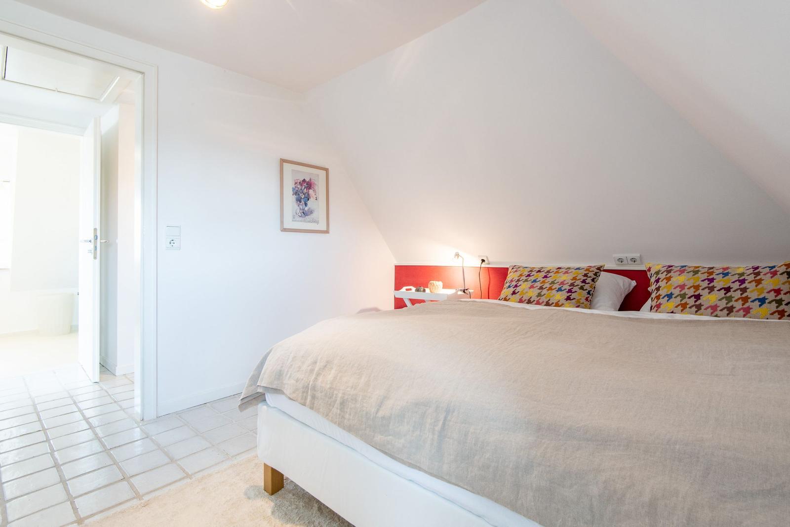 Bedroom with sloped ceiling, bed with colorful pillows, and door to bathroom.