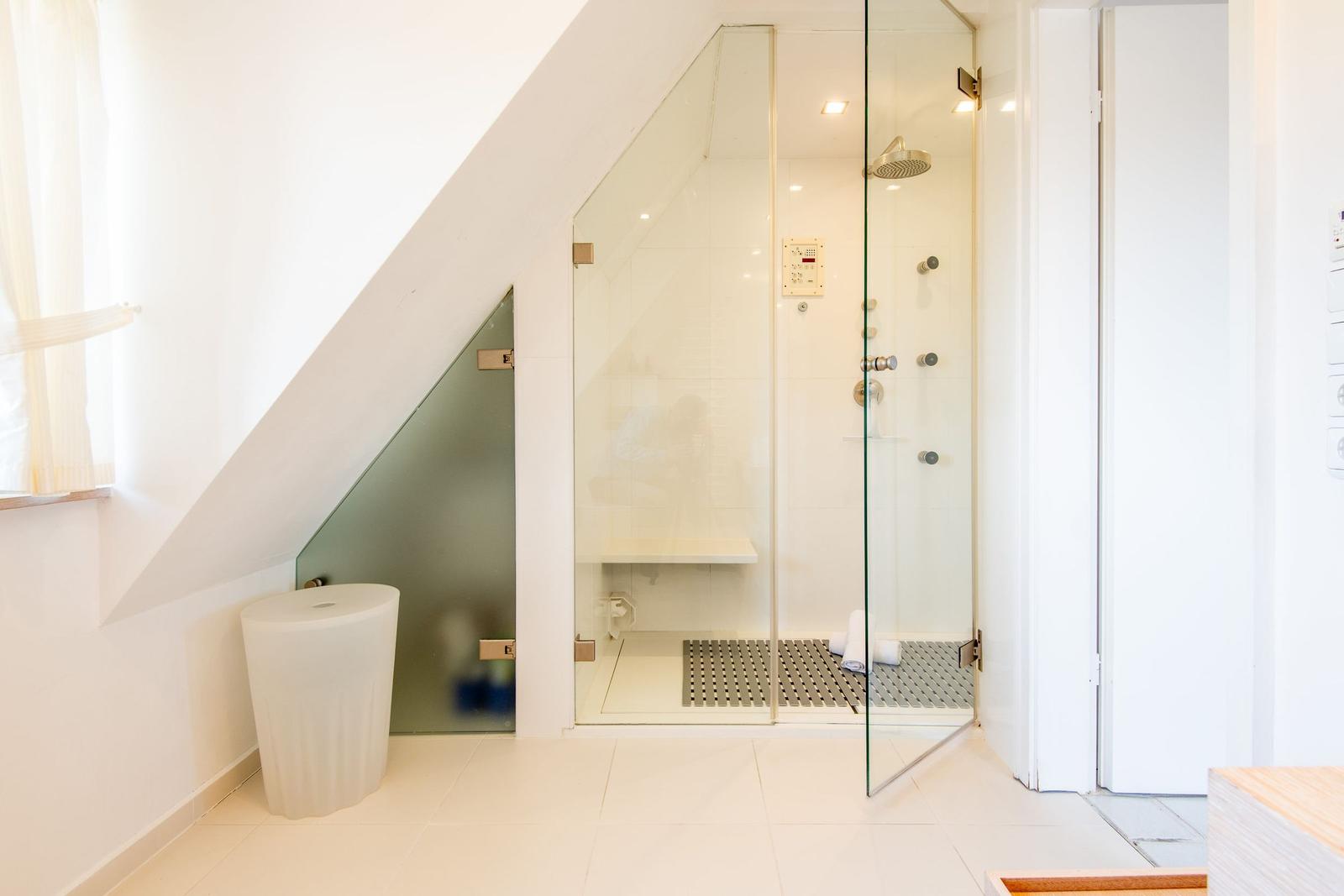 Shower with glass enclosure, toilet, and window under sloped ceiling.