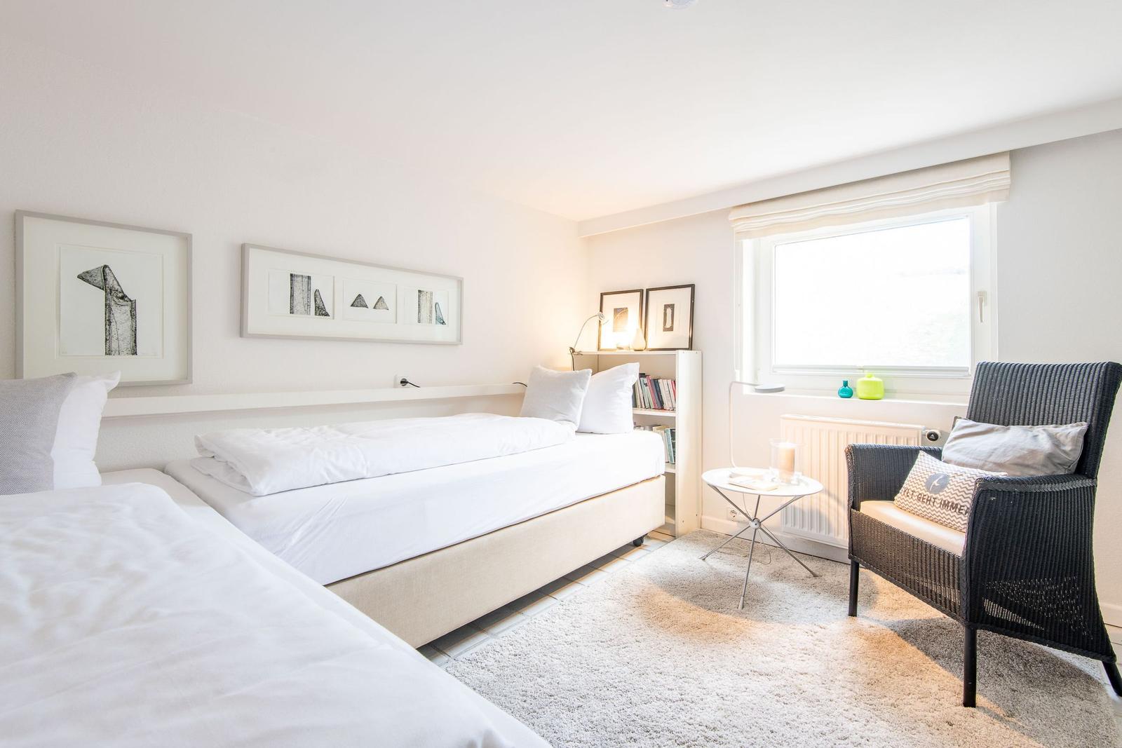 Room with two beds, chair, and window. Wall art and bookshelf.