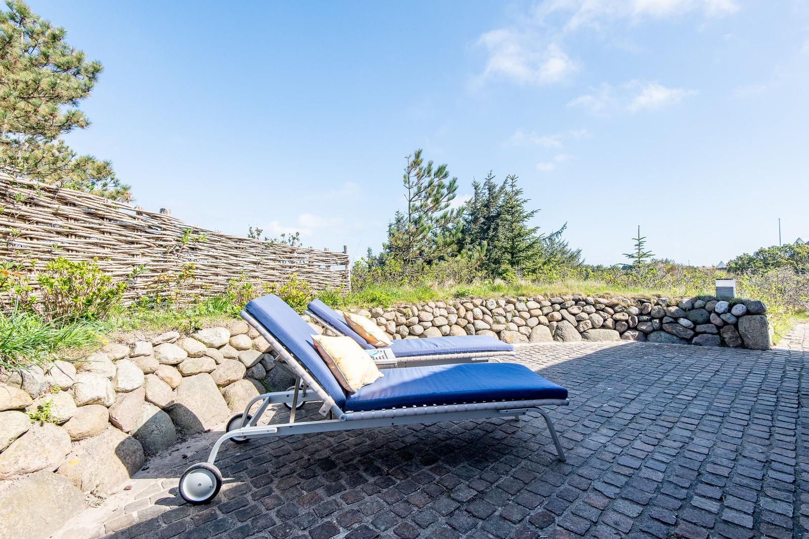 Two blue lounge chairs on cobblestone beside stone wall and wooden fence.