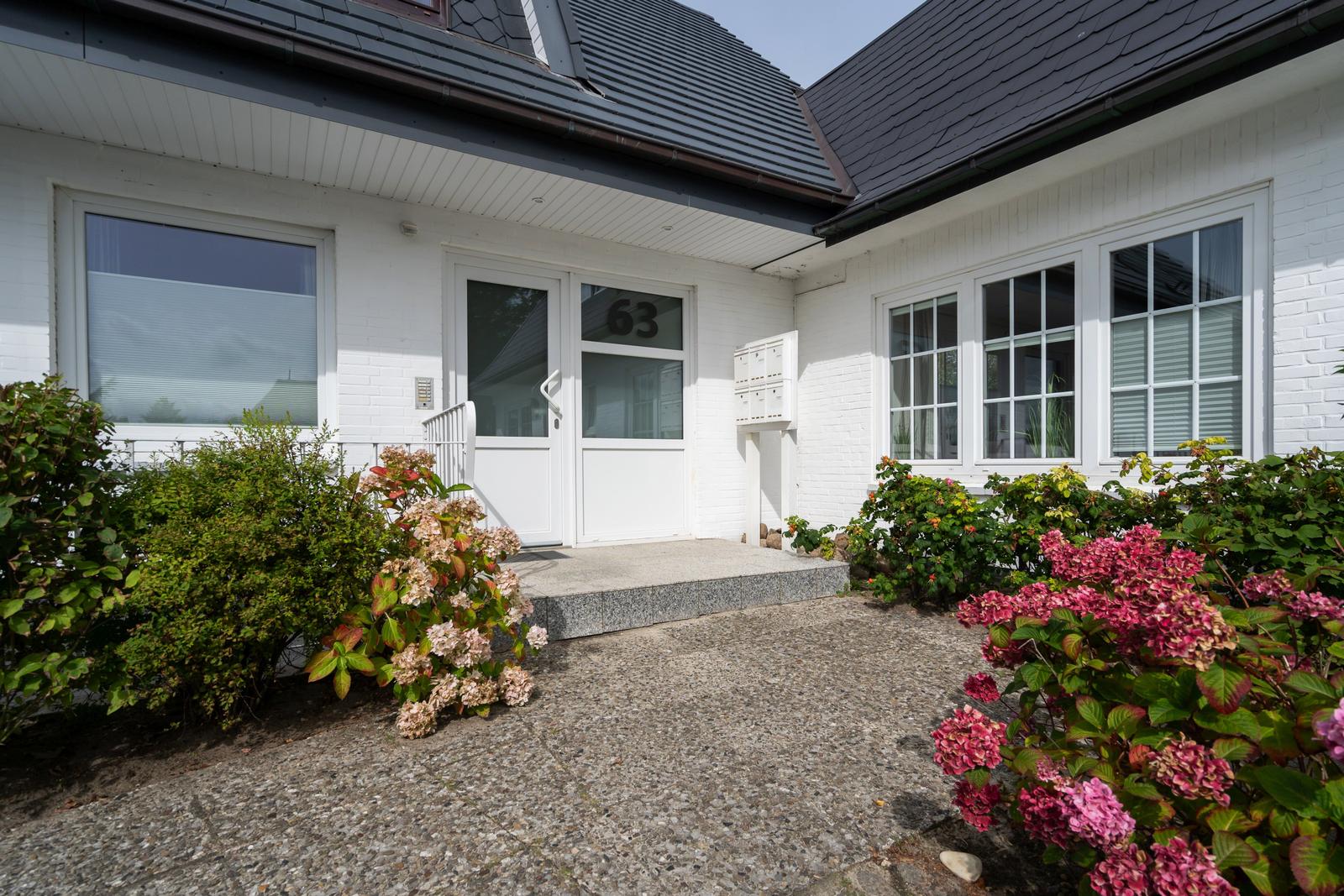 Weiße Villa mit Eingangstür und Blumenbeeten im Vorgarten.
