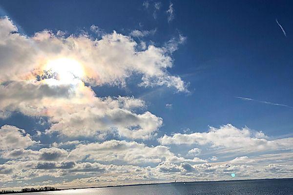 Sonnenlicht durchzieht den Himmel mit weißen Wolken über einem ruhigen Gewässer.