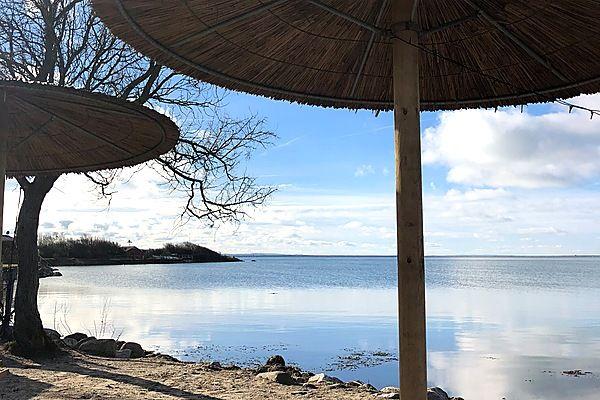 Strand mit Strohschirmen und Blick auf ruhiges Wasser unter blauem Himmel.