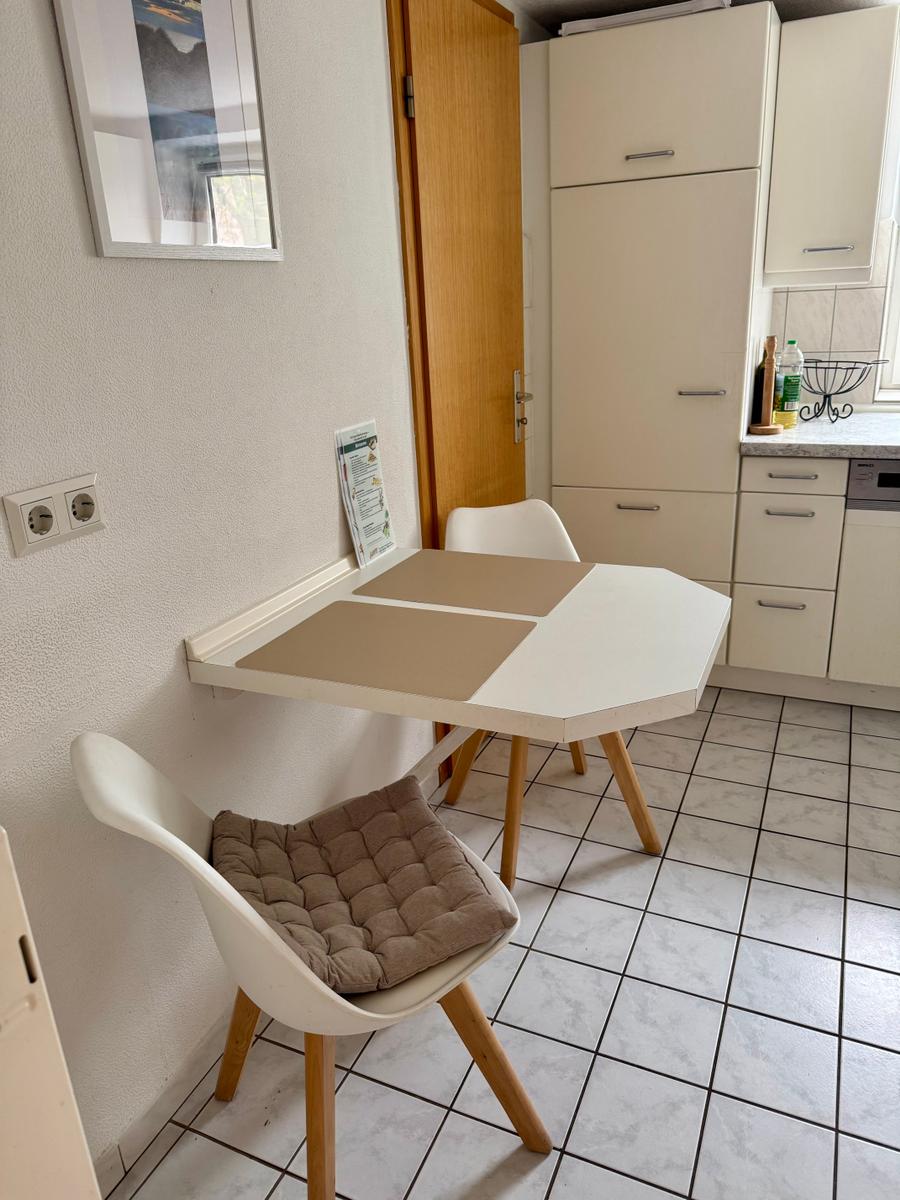 Küchenbereich mit Esstisch und zwei Stühlen. Weiße Schränke und Fliesenboden.