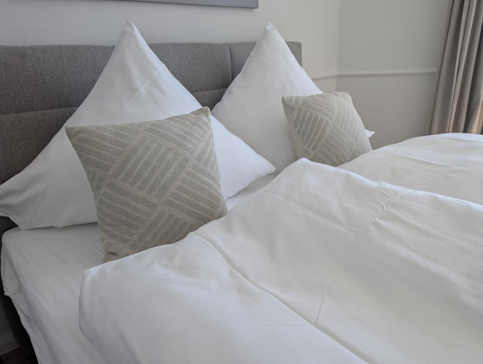 Doppelbett mit weißen Laken und Kissen, grauer Kopfteil, Wand und Vorhang im Hintergrund.
