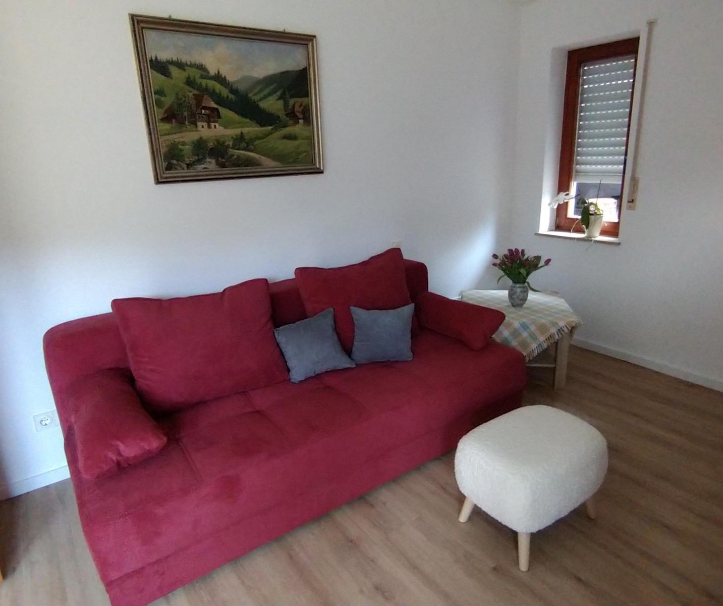 Ein Wohnzimmer mit rotem Sofa, grauen Kissen, weißem Hocker und Wandbild.